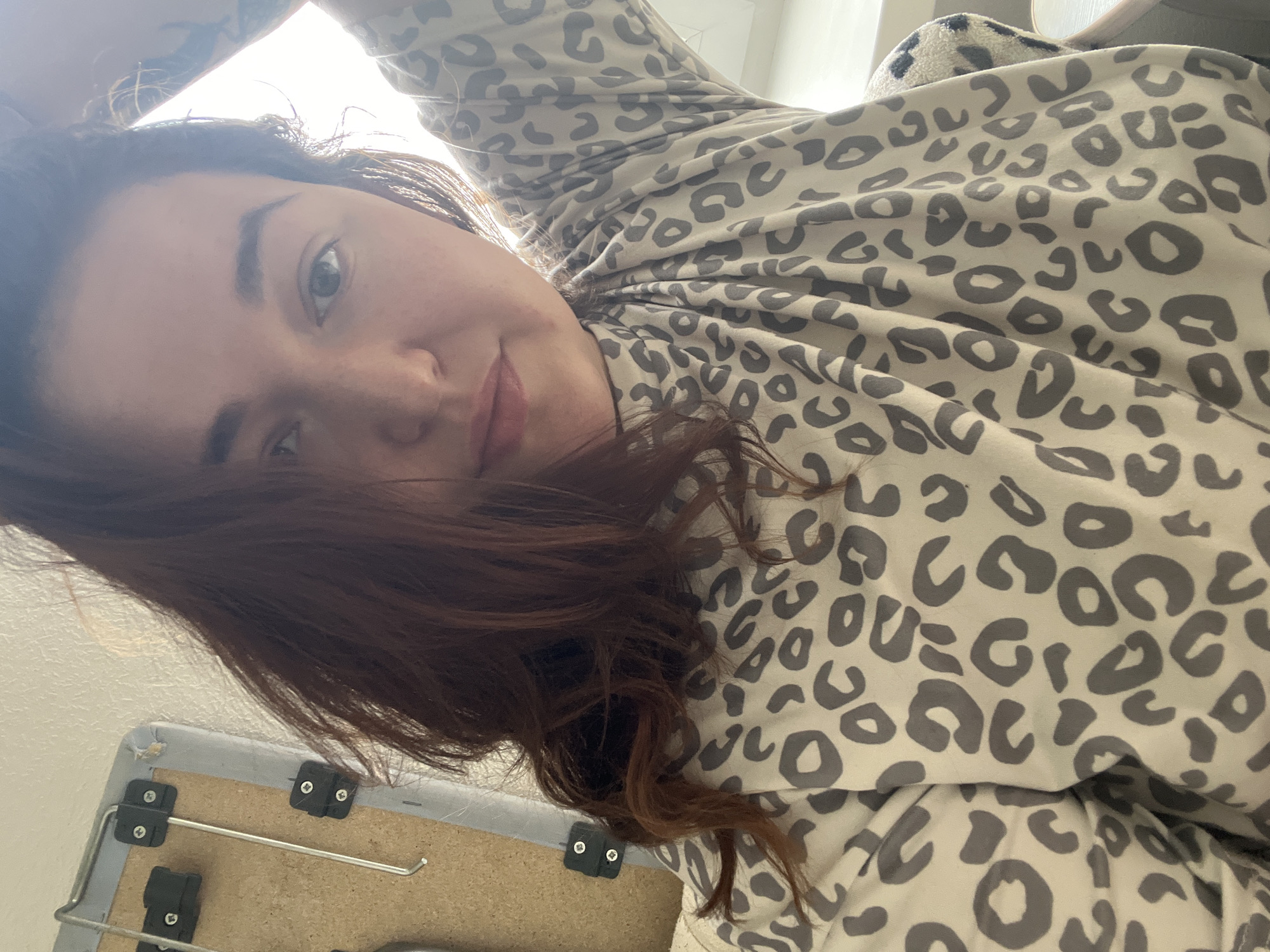 The image shows a selfie of a young woman with auburn hair and a leopard-print shirt. She is indoors, possibly in a bedroom, as evidenced by the presence of what appears to be a bed or couch with a patterned throw. A corkboard with metal attachments is visible in the background, suggesting it might be mounted on a wall. The lighting suggests it is daytime, with natural light filtering in from a window. There are no discernible elements in the image that would allow for the identification of a specific location within Chelyabinsk, Russia.