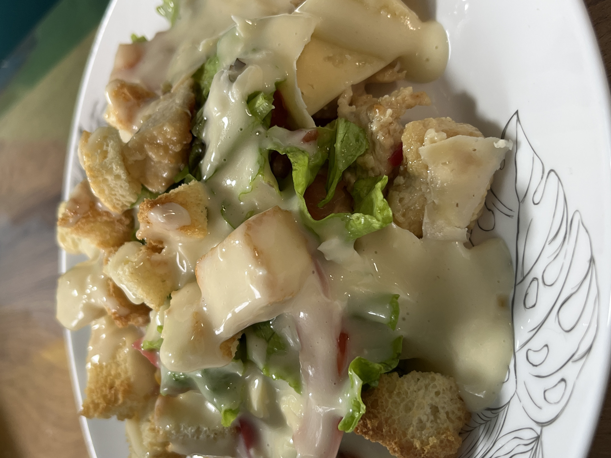 A close-up, overhead shot showcases a plate of Caesar salad. The salad features an abundance of creamy dressing poured over a mix of crisp green lettuce, croutons, and what appears to be chicken pieces and tomatoes. The white plate has a subtle, decorative black line drawing pattern around its rim. The background is blurred, suggesting a shallow depth of field, with hints of a wooden surface and possibly a green object. No specific location in Nizhny Tagil, Russia, can be identified from this image.