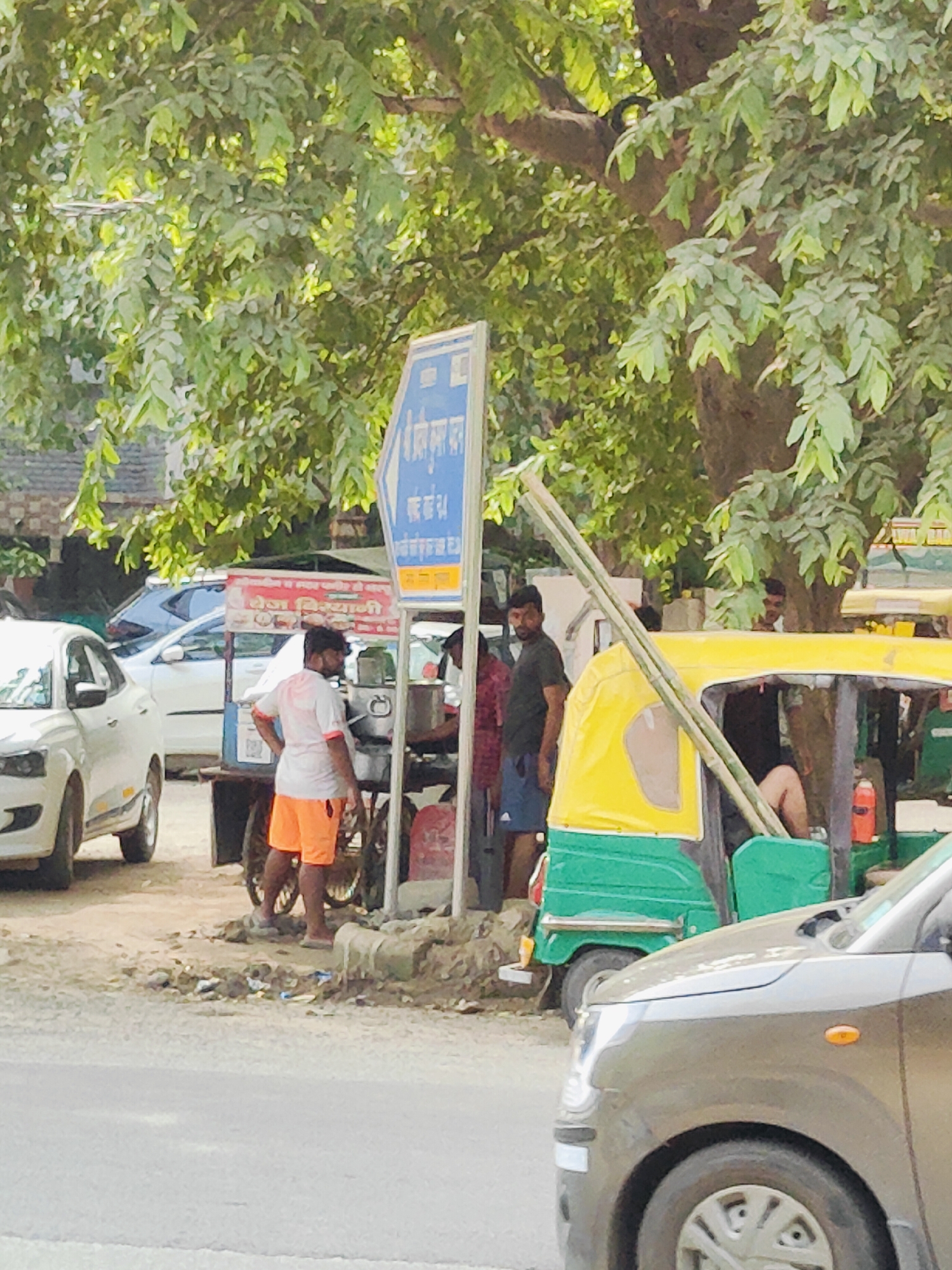 In Gurgaon, India, an outdoor scene depicts a street with several vehicles and people. In the foreground, a grey car is parked. Behind it, a green and yellow auto-rickshaw is visible, with a person sitting inside. To the left of the auto-rickshaw, a man wearing an orange shorts and white t-shirt stands near a food cart. Another man in a black t-shirt is standing to the left of the auto-rickshaw. A white car is parked to the far left of the scene. A blue and white sign is visible above the food cart. Tall trees with green leaves provide shade over the area. The ground is covered with dirt and debris.