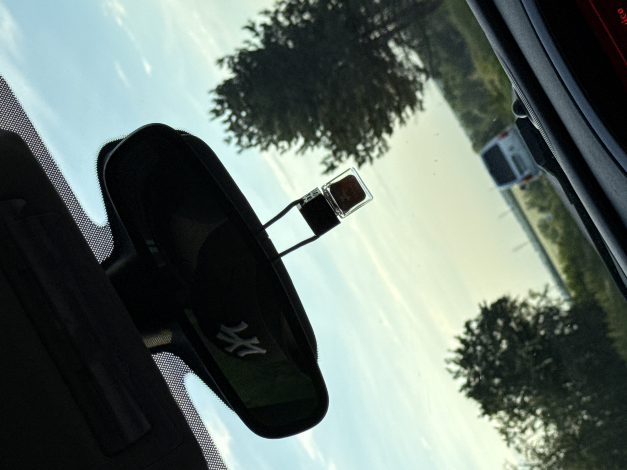 The image shows a view through the windshield of a car, likely a Maserati given the logo on the rearview mirror. The sky is visible through the windshield, along with trees and a road. A small vehicle is visible in the distance on the road. An air freshener is attached to the rearview mirror. The location is unspecified and unidentifiable as Pleven, Bulgaria.