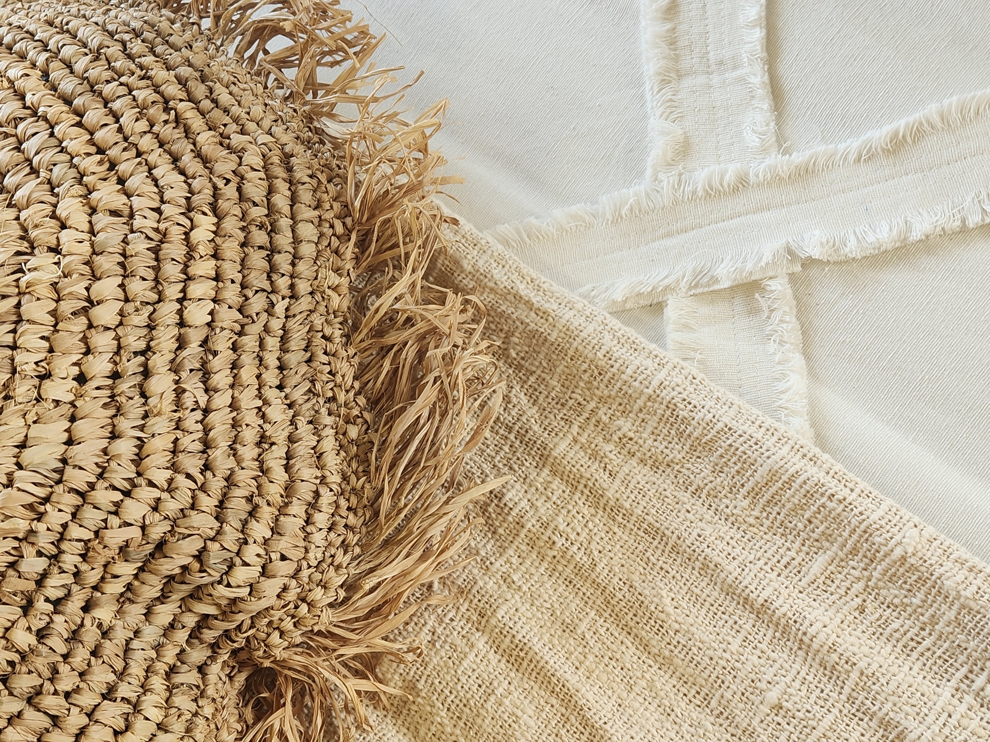 This media file is a close-up, overhead shot of woven natural materials. The main subject on the left is a textured object made of tightly woven straw or raffia, with a fringe of loose straw strands along its edge. This object appears to be a decorative item or part of a larger piece, like a basket or hat.
To the right of the straw object, there are two layers of fabric. The bottom layer is a coarse, loosely woven natural fabric in a light beige color, showing a clear woven texture. Overlapping this is a lighter, off-white fabric with a more uniform weave, characterized by distinctive horizontal bands of frayed threads along its edges. The arrangement suggests a still life composition, likely staged for a photograph.
The lighting is soft and diffused, indicating it could be natural light from a window or a studio setting. The overall aesthetic is organic, rustic, and minimalist, with a focus on texture and natural materials. There are no people visible, nor any specific clues about the time of day, weather, or precise location beyond the general context of Maitland, USA. The composition is abstract due to the close-up perspective, with no visible text. The scene evokes a sense of calm and natural beauty.