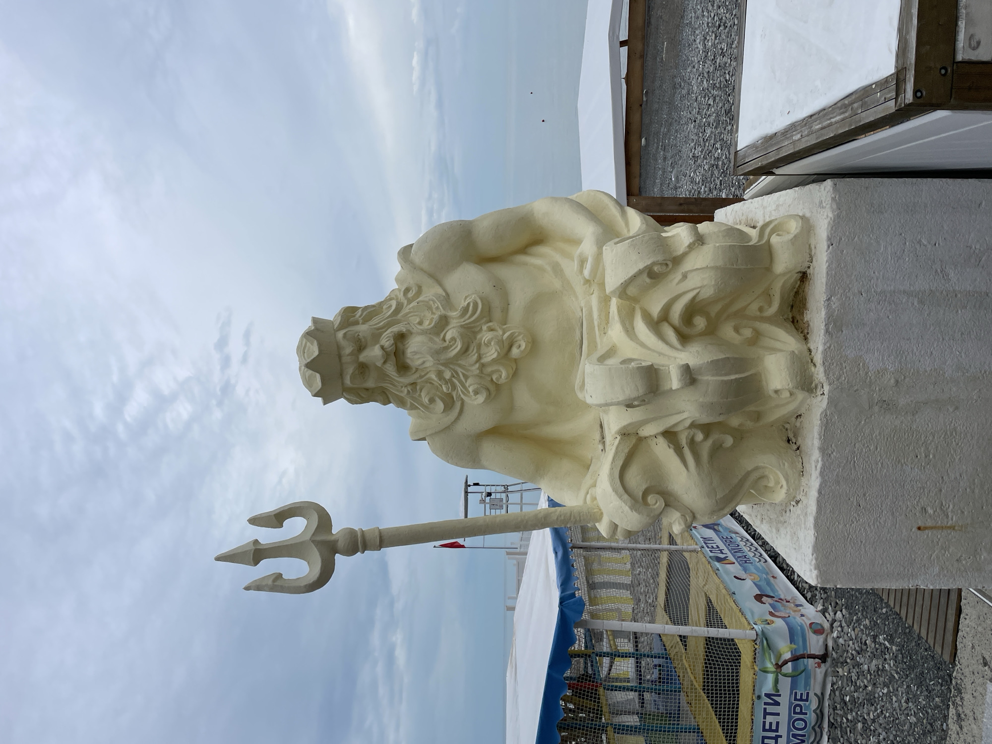 The image depicts a sculpture of Poseidon, the Greek god of the sea, holding a trident. The sculpture is made of a light-colored material and is positioned outdoors. The background includes the sea, a cloudy sky, and what appears to be a beach area. A sign in Russian is visible near the base of the sculpture. The sculpture's location is the city of Sochi, Russia. A beach-like environment is indicated by the pebbles and the marine theme.