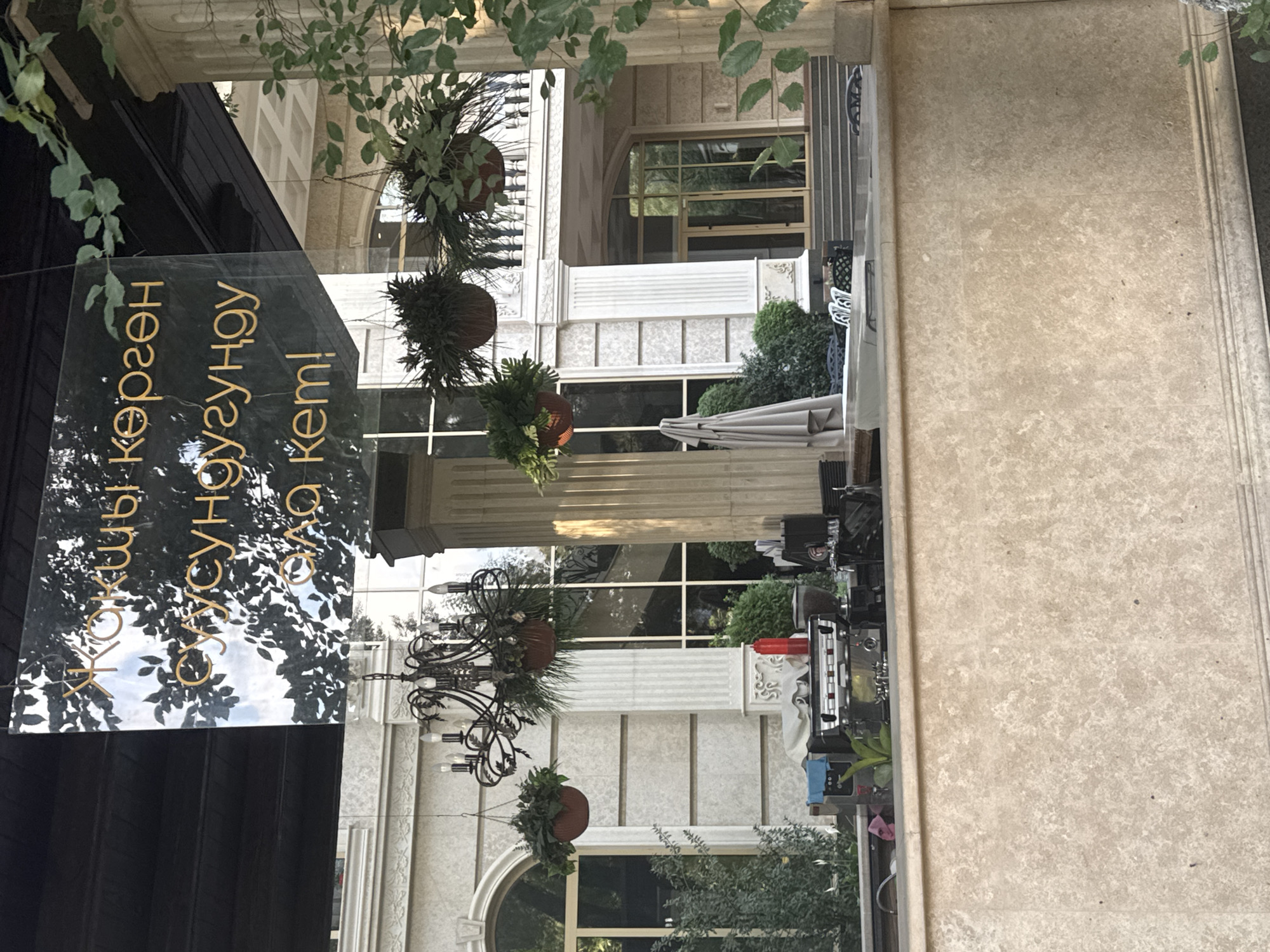 The image shows a sunny outdoor scene in front of a building in Bishkek, Kyrgyzstan. A sign with Cyrillic text hangs from the ceiling. In the foreground, a marble-like counter is visible, with a coffee machine and other bar equipment on top. Behind the counter, there are plants and a chandelier. The building in the background is decorated with columns and arched windows.