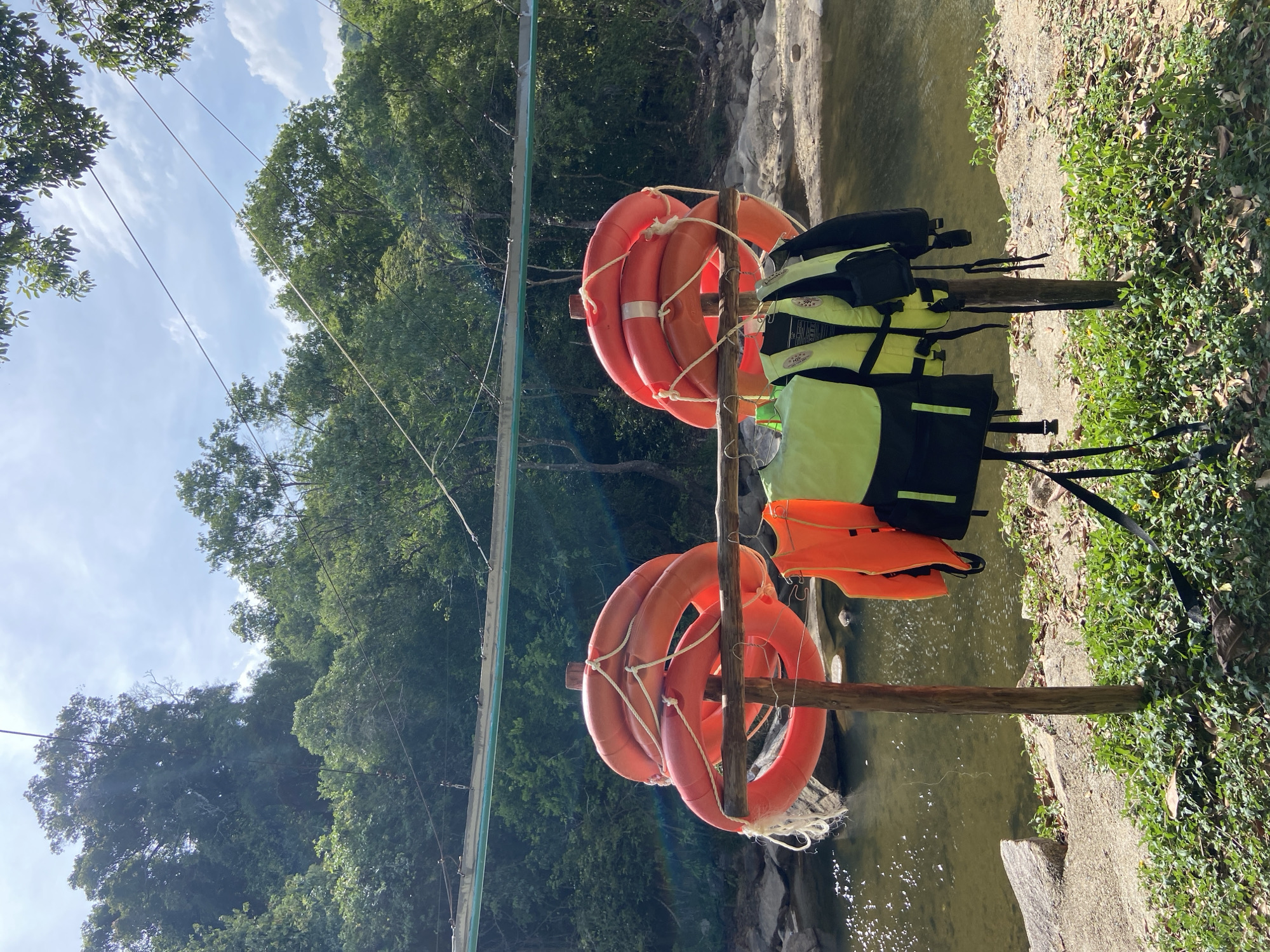 The image shows a riverbank scene with safety equipment. In the foreground, two wooden posts support a horizontal bar from which several bright orange and lime green life vests are hanging. Behind the posts, a stack of orange life preservers is suspended from a horizontal beam. A shallow river flows behind the equipment, with rocky banks and lush green trees lining the opposite side. Overhead, a cable is strung across the scene, possibly part of a zipline or suspension bridge. The sky is partly cloudy. The scene appears to be a recreational area, likely a river for activities like kayaking or tubing, located in a natural, forested environment. There are no people visible in the image. The image context provided states the location is Hoa Thang Ward, Vietnam.