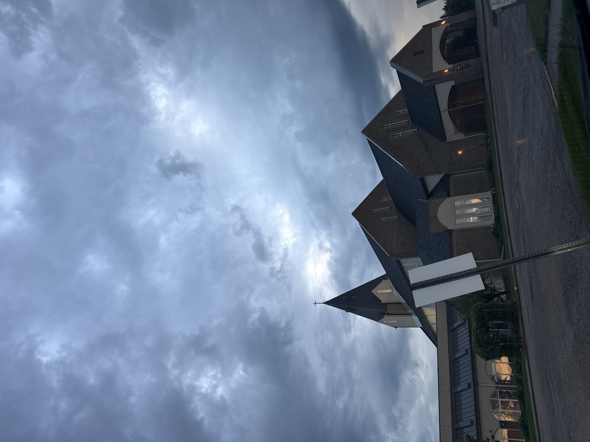 A church building with a tall steeple and a brick facade is shown under a cloudy, grey sky. The church appears to be a modern structure with large windows. In the foreground, there is a street with some puddles, suggesting recent rain. A blank white sign on a pole is visible to the right of the church. The image was taken in Stony Plain, Canada.