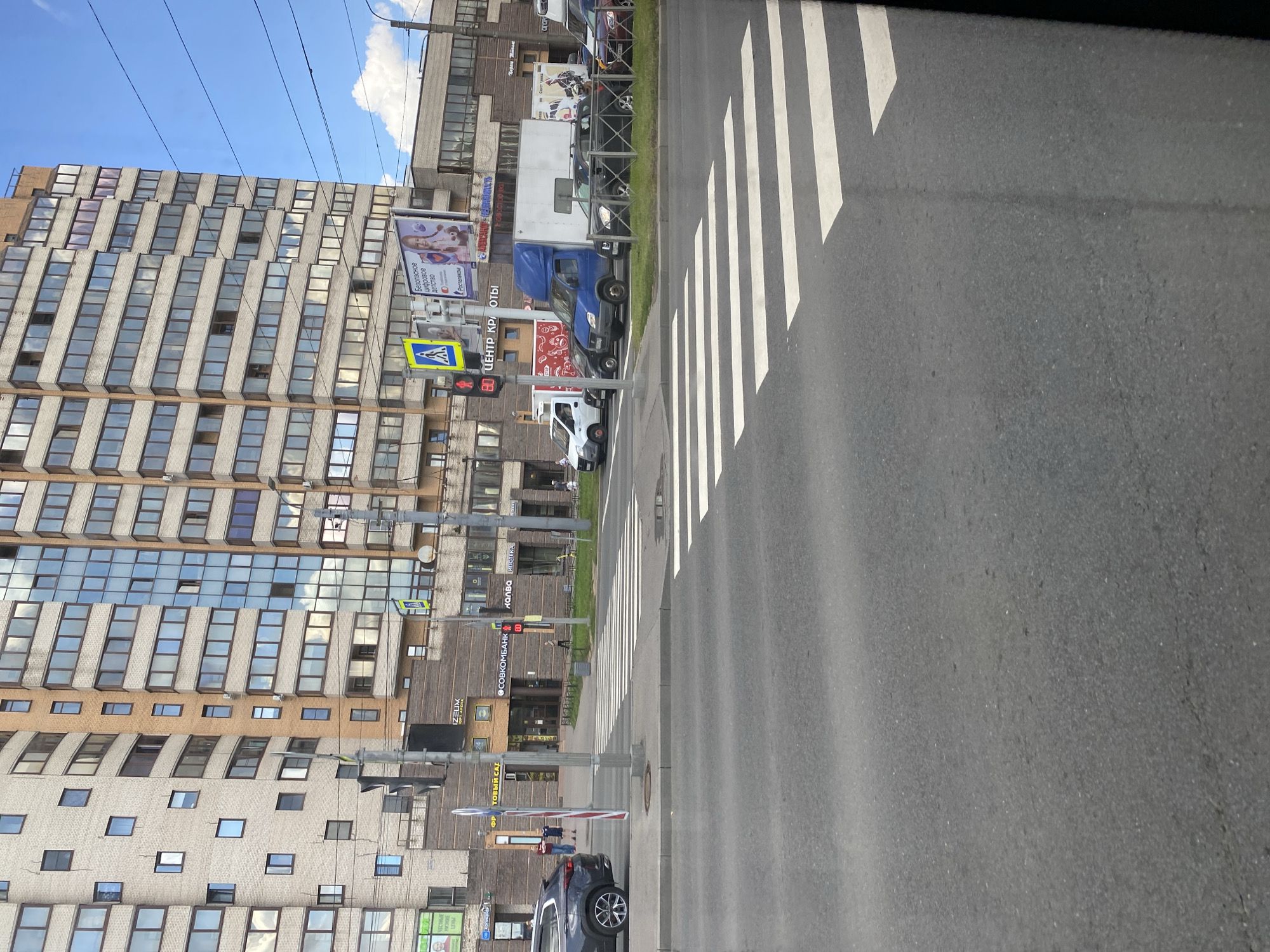 The image depicts a street scene in Saint Petersburg, Russia, focusing on a pedestrian crossing in front of a modern apartment building. The building, with its beige brick facade and numerous windows reflecting the blue sky and clouds, dominates the background. Commercial establishments with signage are visible at the base of the building, including "IZELUX," "ФРУКТОВЫЙ САД" (Fruit Garden), and "СОБКОМБАНК" (Sobcombank). A blue delivery truck, a white van, and several cars are parked or driving near the crossing. Pedestrian traffic appears minimal, with a few individuals visible walking near the building. Traffic lights and street signs, including a pedestrian crossing sign, are also present. The foreground shows the asphalt road and the white stripes of the pedestrian crossing. The overall environment suggests a typical urban setting on a clear day.