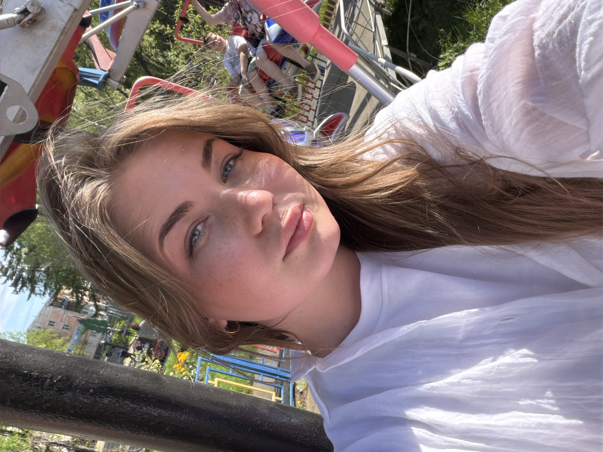 A young woman with light brown hair and fair skin is taking a selfie. She has light-colored eyes and freckles. She is wearing a white t-shirt. The background shows a carousel ride with people on it. Behind the ride is a park with buildings and trees. Based on available information, the location is likely in the city of Chaikovsky, Russia.