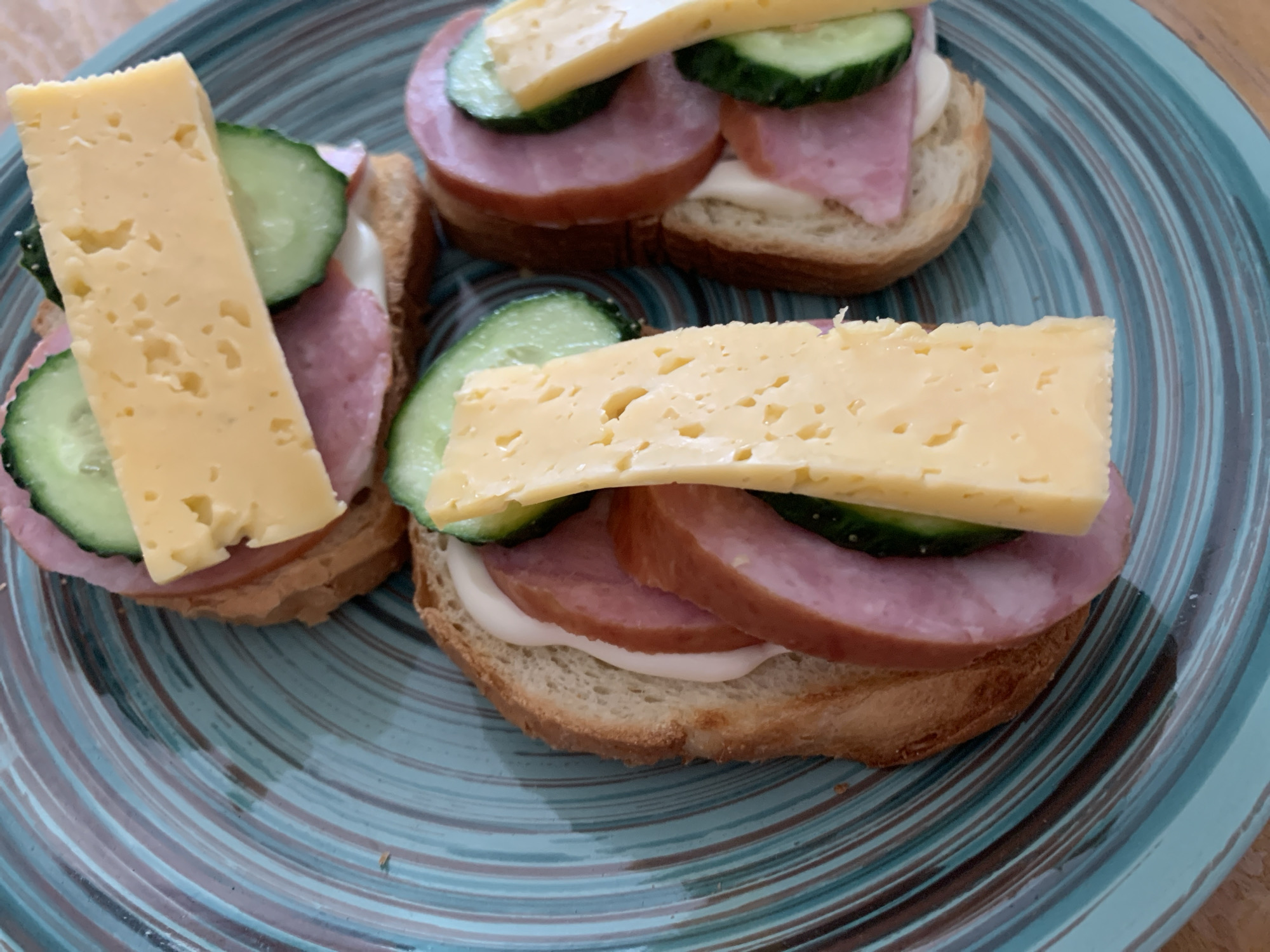 A close-up, overhead shot shows three open-faced sandwiches on a blue, swirled ceramic plate. Each sandwich is made with a slice of toasted bread, topped with a dollop of white spread (likely mayonnaise), several slices of pink, processed sausage, and slices of green cucumber. A thick slice of yellow cheese crowns each sandwich. The sandwiches are arranged in a diagonal line across the plate, with the plate resting on a wooden surface. The scene is consistent with a casual meal prepared and photographed in a kitchen or dining area within a residential setting, possibly in Stavropol, Russia, given the user's prompt. There are no people or animals visible in the image. The lighting appears to be natural, possibly from a nearby window. The focus is sharp on the sandwiches, highlighting their textures and colors.