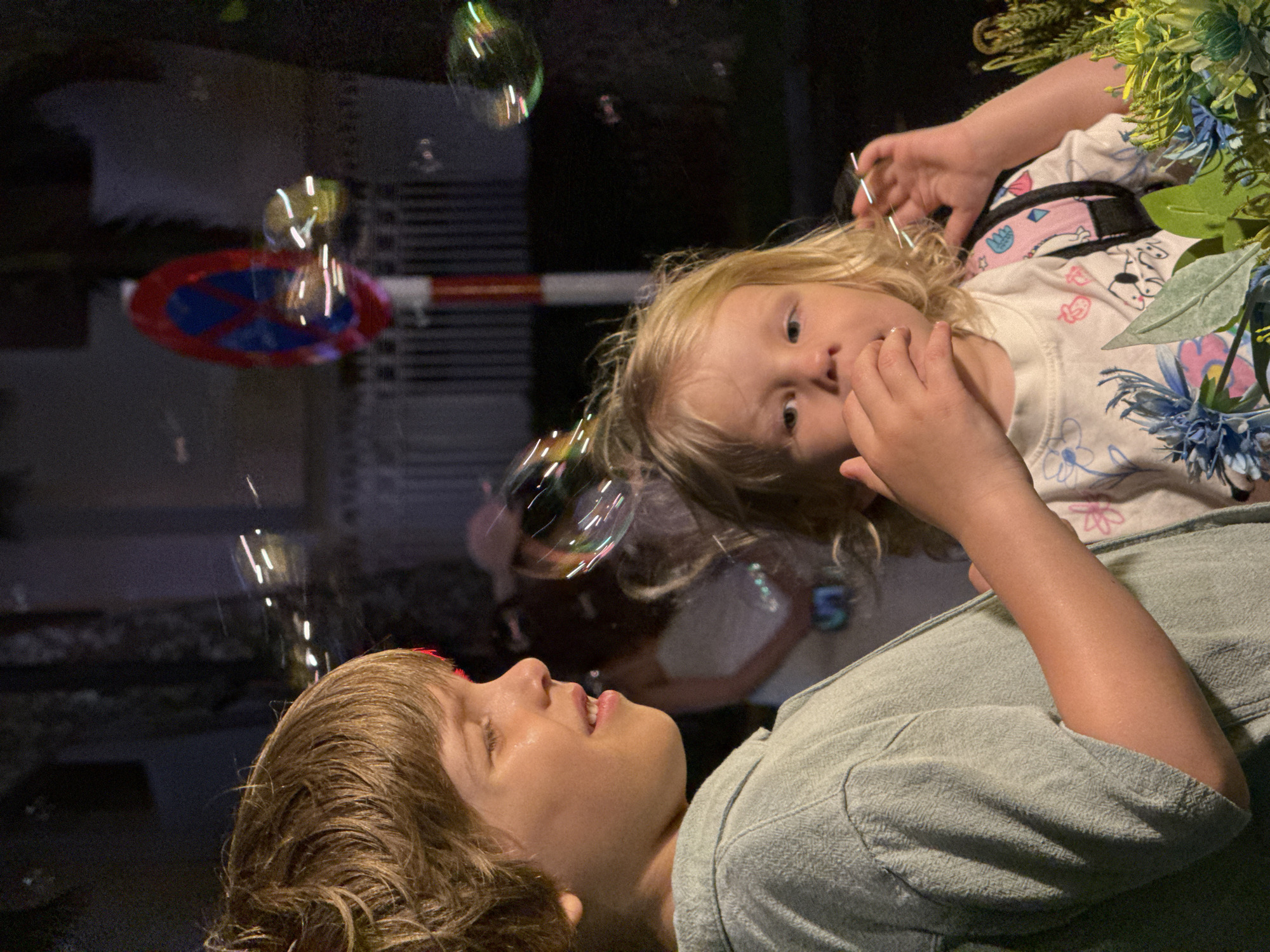 This intimate portrait, taken at night in Nha Trang Ward, Vietnam, captures a playful moment between two children, surrounded by shimmering bubbles. The image is oriented so the subjects appear to be lying down, seen from above.
In the foreground, an older child, likely a boy with medium brown hair, lies on his back, looking upwards with his mouth slightly open, possibly blowing or reacting to the bubbles. He wears a light green or grey casual top. His expression is one of calm focus or perhaps wonder.
Above him, a younger child with blonde hair looks directly out of the frame with wide, curious eyes. She is wearing a white t-shirt adorned with colorful patterns and cartoon figures, along with blue and pink floral designs. Her left hand is raised near her face. To her right, a lush arrangement of green and blue foliage provides a natural frame.
Numerous iridescent bubbles float throughout the dark background, reflecting ambient lights and creating bright streaks. In the blurred background, an urban environment is visible: a "No Stopping" or "No Parking" street sign (a blue circle with a red border and diagonal slash) is prominent, alongside a horizontal red and white barrier and the dark, textured facade of a building. A faint reflection of another person, possibly an adult, can be seen in the bubbles or background, suggesting an adult's presence facilitating the bubble-blowing activity. The low light conditions emphasize the glowing quality of the bubbles and the children's expressions of engagement and wonder. No explicit text is visible.