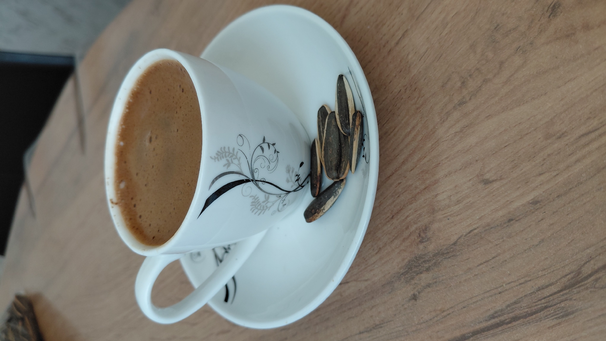 A close-up, eye-level shot captures a white coffee cup filled with dark, frothy coffee, resting on a matching saucer. Several sunflower seeds are scattered on the saucer in front of the cup. The cup and saucer are adorned with a delicate black and grey floral pattern. The scene is set against a backdrop of a wooden table with a prominent grain pattern. The lighting is soft, casting subtle shadows. The image was taken in Abovyan, Armenia.