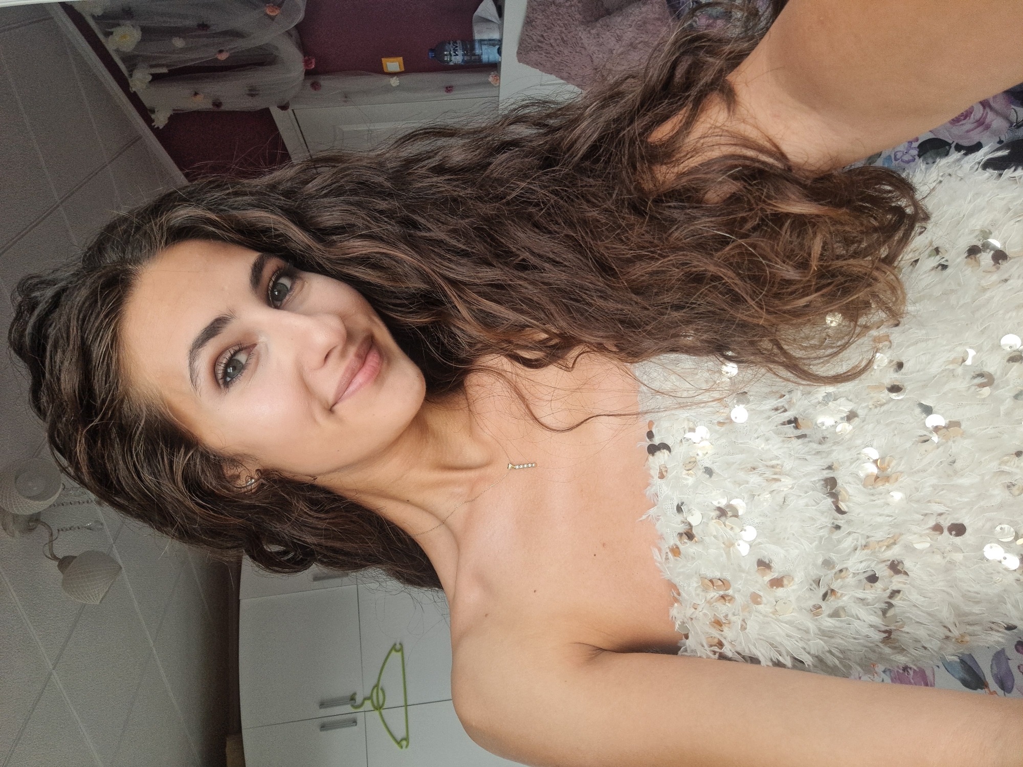 A young woman with long, wavy brown hair is pictured from the chest up, facing the camera with a slight smile. She is wearing a strapless white top adorned with white feathers and scattered sequins. A delicate gold necklace with a single pendant hangs around her neck. Her skin is smooth and lightly tanned.
The background appears to be an indoor setting, possibly a bedroom. To the left, a white closet with handles is visible, with a green hanger hanging on one of the doors. Above, a white ceiling with light fixtures is present. To the right, a partially visible dark purple wall can be seen, decorated with sheer white fabric draped with small pink and white flowers. A shelf holds a bottle of water and some other items. The overall impression is one of a personal, domestic space.