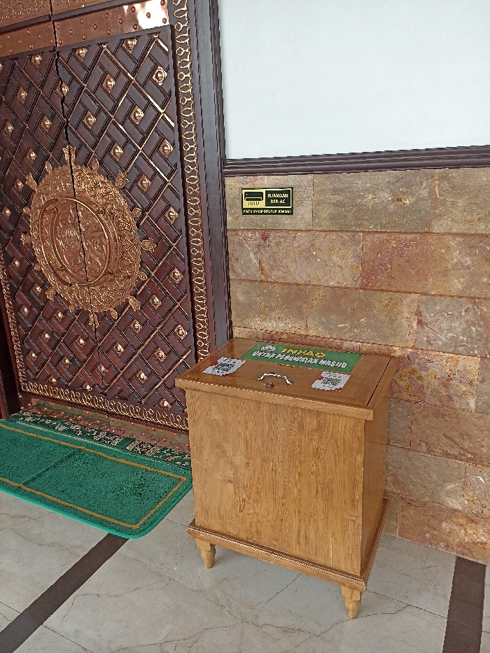 The image captures a static scene at what appears to be the entrance of a building, likely a mosque, given the context clues. No people are visible.

The main subjects are an intricately carved wooden door and a wooden donation box positioned in front of a stone-clad wall. The door is dark brown, adorned with elaborate gold-colored metallic patterns, including geometric designs and a prominent circular medallion in the center. A green mat with a gold border lies on the light-colored tiled floor directly in front of the door.

To the right of the door, a plain, light-brown wooden donation box with decorative legs stands on the tiled floor. Its top surface features a green area with white text, reading "INFAQ UNTUK PEMBANGUNAN MASJID," which translates to "DONATION FOR MOSQUE CONSTRUCTION." There are also several QR codes visible on the top of the box, likely for electronic donations, though the specific details on them are not clearly legible.

Behind the donation box and to the right of the door, a wall is covered with natural stone cladding, exhibiting varied shades of brown, orange, and grey. A small, black rectangular sign is affixed to this wall. The text on the sign, in white, states: "RUANGAN BER-AC" (Air-conditioned Room) and "PINTU HARAP DITUTUP KEMBALI" (Please Close the Door Again).

The setting suggests an interior or sheltered exterior area of a public building, possibly in Plemahan, Indonesia, as indicated by the language on the signs and donation box. The lighting appears to be ambient, indicative of daytime.