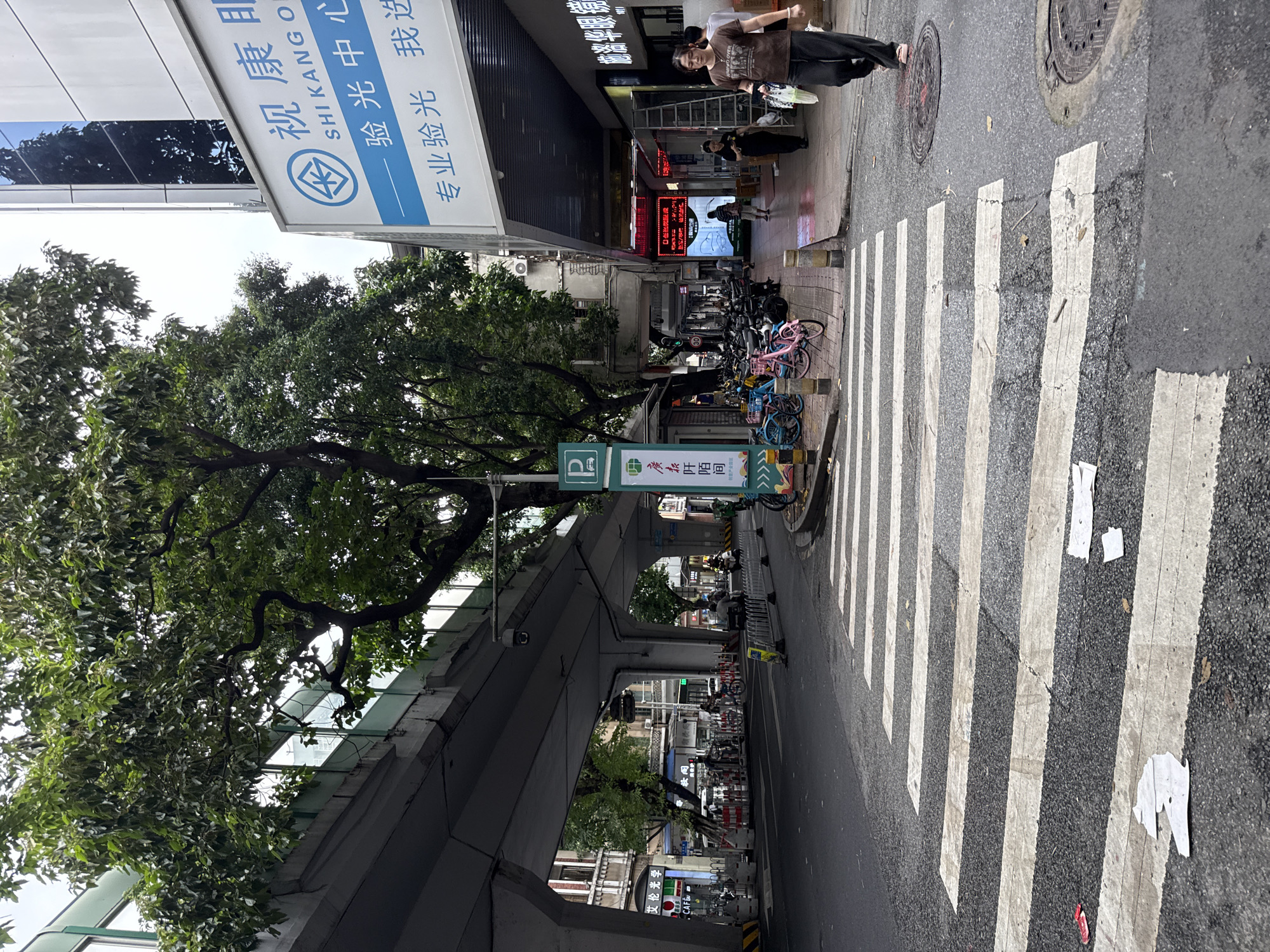 The image captures a street scene in Yuexiu District, China, with a pedestrian crosswalk prominently in the foreground. An elevated roadway or overpass dominates the upper portion of the frame, casting a shadow over the street. To the right, a building facade features a large sign with Chinese characters and the text "SHI KANG O". A woman walks across the crosswalk from right to left, carrying plastic bags. To the left of the crosswalk, parked bicycles are visible on the sidewalk, and further down the street, more businesses and a traffic light can be seen. The environment appears to be an urban setting with moderate pedestrian and vehicular activity.