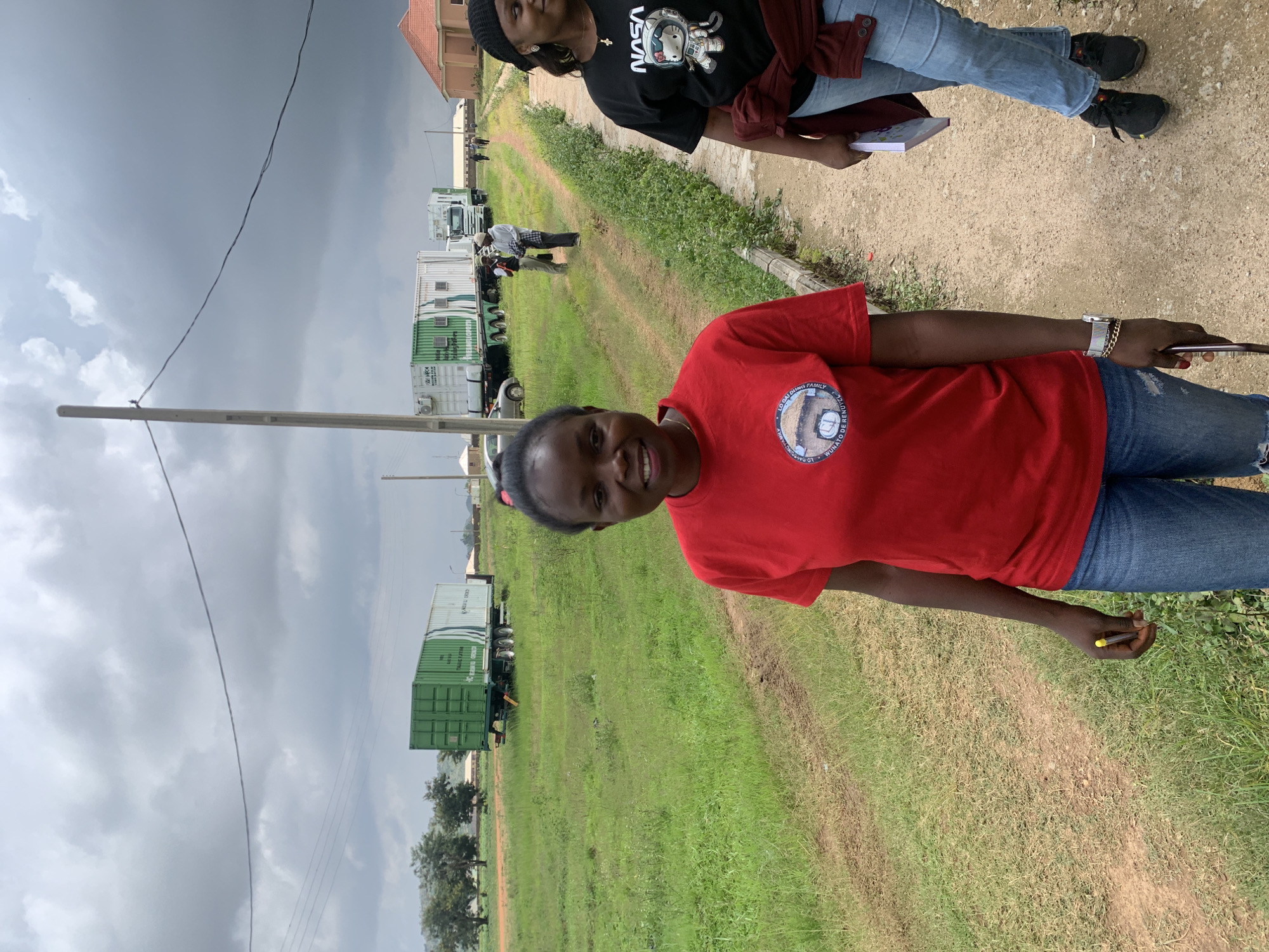 In Gwafan, Nigeria, two women stand on a dirt path in front of a row of shipping containers on trailers, under a cloudy sky. The woman in the foreground, wearing a red t-shirt and blue jeans, smiles at the camera. She holds a pen in her right hand and wears a watch and bracelet on her left wrist. To her right, a second woman, wearing a black t-shirt with a NASA logo featuring a cat astronaut, jeans, and a black beanie, looks to the right. She holds a book in her left hand. In the background, several green shipping containers are visible, some on trailers pulled by trucks. A few other people are walking further down the path. The environment is a grassy area with scattered trees and some buildings in the distance.