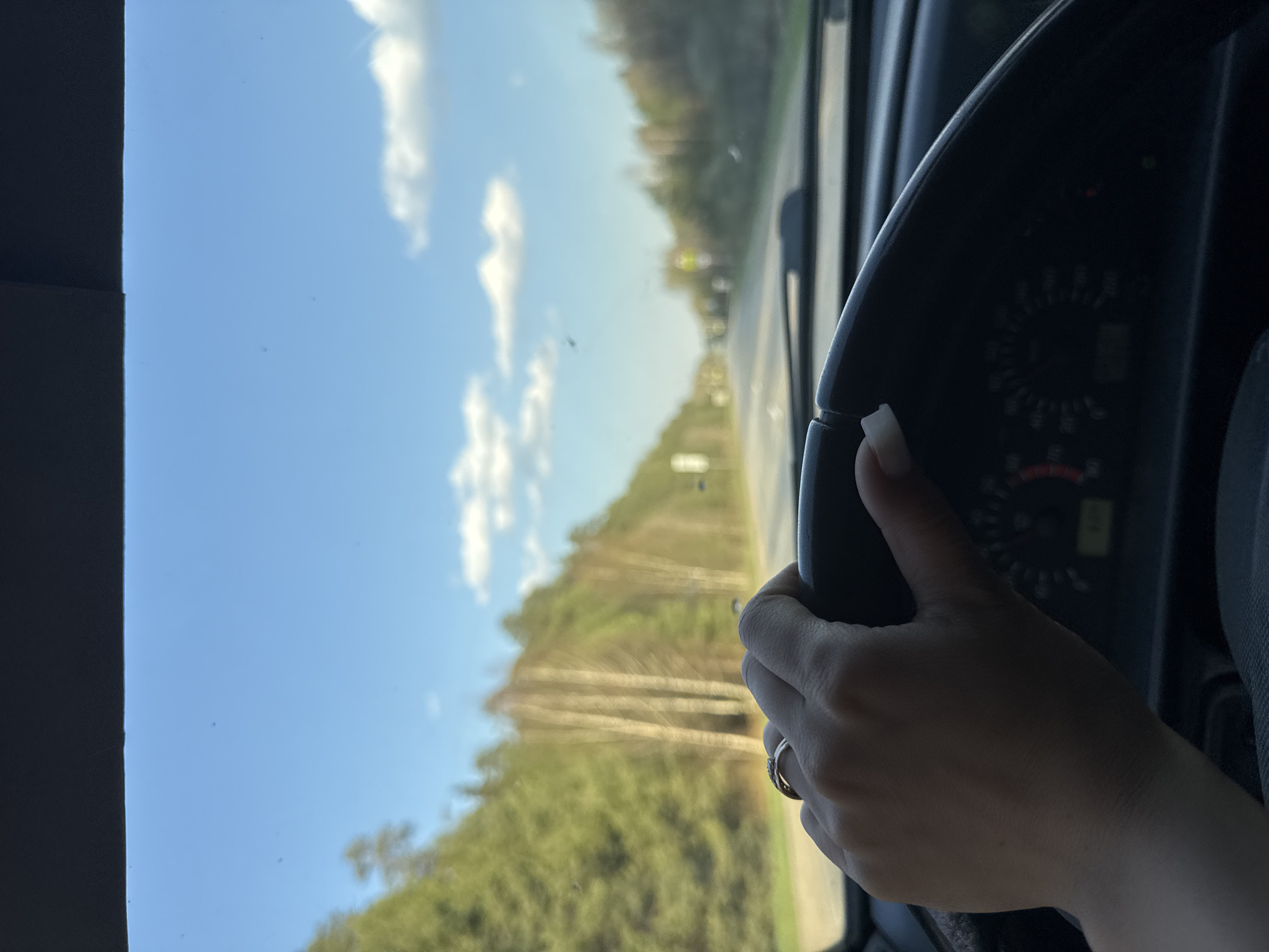 A view from inside a vehicle looking out through the windshield. A person's hand is visible on the steering wheel. The view through the windshield shows a road, trees, and a blue sky with clouds. The location is Chaikovsky, Russia.