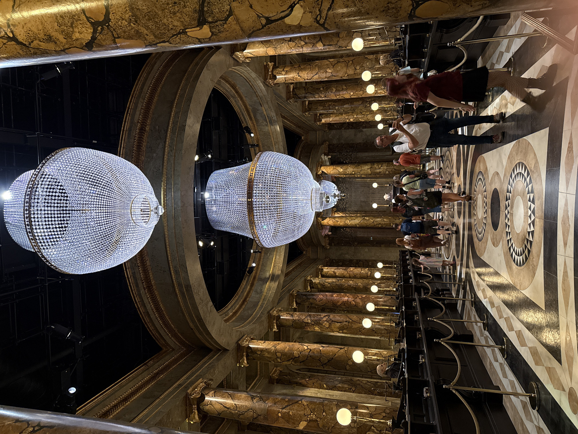 The image depicts the interior of Gringotts Wizarding Bank, a popular attraction at the Warner Bros. Studio Tour London – The Making of Harry Potter, located in Leavesden, near Watford, Hertfordshire, United Kingdom. The scene is characterized by its opulent and grand architecture, reminiscent of a bank's main hall. Two massive, ornate crystal chandeliers hang from the high, dark ceiling, illuminating the space. The hall is lined with tall, marble-effect columns, predominantly in shades of gold and brown, with intricate gold detailing on their capitals and the arched ceilings above.
In the foreground, several people are visible. A man in a white t-shirt and jeans is taking a photo with his phone, facing towards the interior. To his right, a woman with red hair and a black skirt is walking away from the camera. Further down the hall, a group of people, including children and adults, are walking towards the left. They appear to be visitors exploring the attraction.
The floor is adorned with a striking patterned mosaic of geometric shapes, primarily in black, white, and beige, with gold accents. Along the sides of the hall are long, dark counters, resembling bank teller stations, behind which several figures are seated, presumably staff members of the attraction. Brass ropes on stanchions create queue lines for visitors. Small, globe-shaped lamps on tall stands are placed along the counters, adding to the ambient lighting. The overall impression is one of a meticulously designed, immersive environment that recreates the magical world of Harry Potter for visitors.
