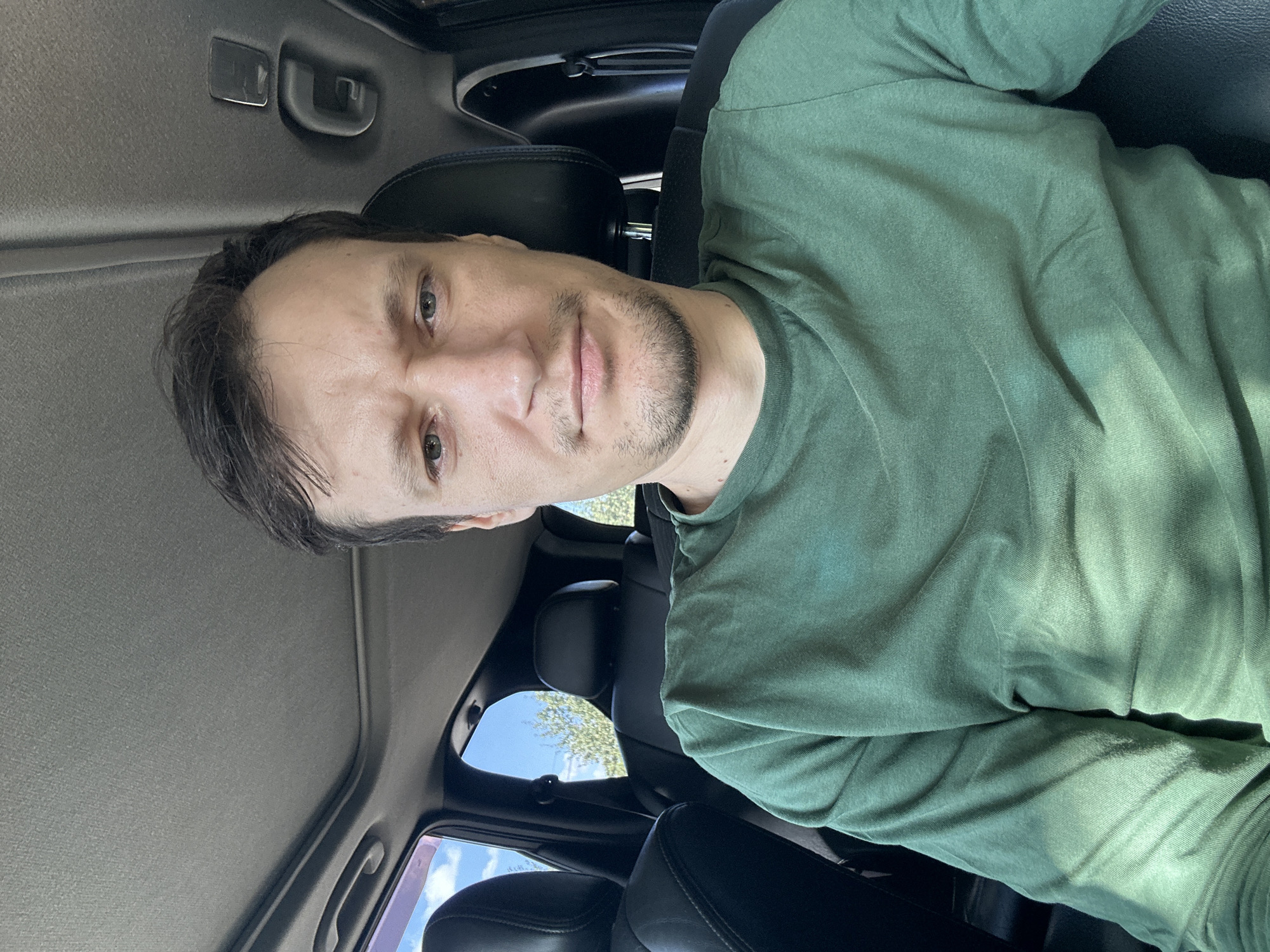 A man, appearing to be of European descent, is seated in the passenger seat of a car. He is looking directly at the camera. The interior of the car is visible, including black leather seats, the car's headliner, and a sunroof. Through the windows, trees and a bright blue sky with some clouds can be seen, suggesting an outdoor setting. The man is wearing a long-sleeved green t-shirt. The image does not provide specific information about the city of Makiv, Ukraine.