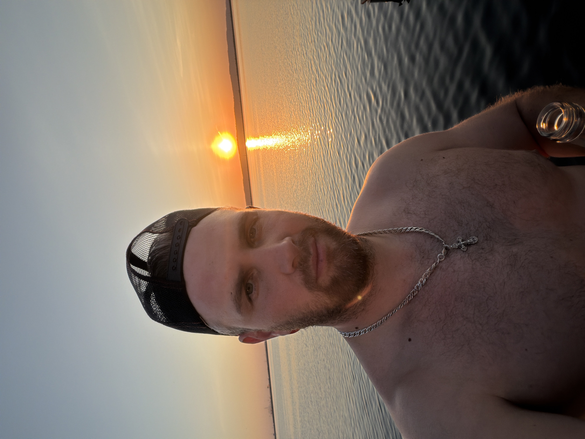 A medium shot captures a fair-skinned man with a beard and a black mesh baseball cap, set against a sunset over a body of water. The man is shirtless, wearing a silver chain with a cross pendant. He is holding a clear glass bottle in his right hand. The water reflects the orange and yellow hues of the setting sun. The image was taken in the city of Chaikovsky, Russia.