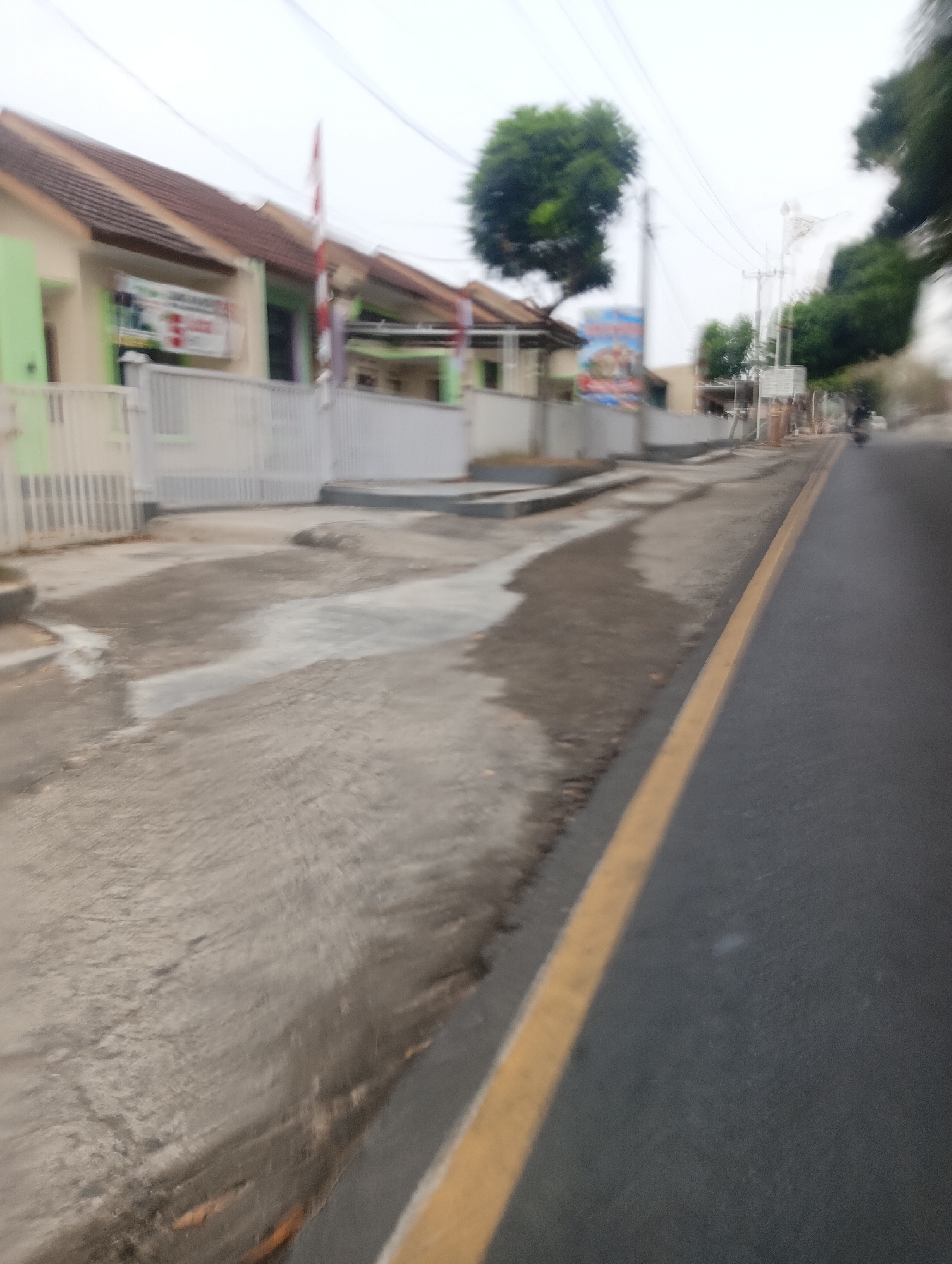 The image depicts a blurry street scene in Mekarmukti, Indonesia, likely captured from a moving vehicle. On the left side of the frame, there are several light-colored houses with brown roofs, characteristic of residential housing in the area. One house has a green facade and a white fence in front. A banner with red and white elements is visible on the front of one of the houses. A flagpole with a red and white flag is also present.
The street itself is paved with asphalt and has a yellow line marking the edge of the lane. The area beside the road appears to be a drainage ditch or a poorly maintained sidewalk with patches of concrete and dirt. A few trees, some appearing lush and green, are scattered throughout the background. In the distance, utility poles and power lines are visible. A motorcycle is present on the right side of the road, suggesting traffic in the area. The overall impression is one of a somewhat modest and developing urban or suburban environment. The lighting suggests an overcast or cloudy day.