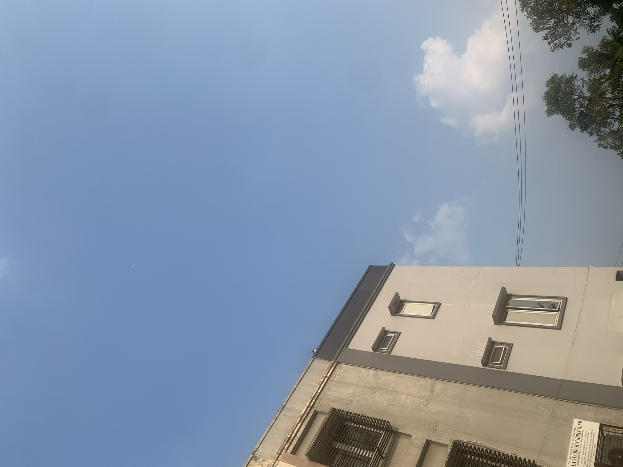 The image depicts a section of a building against a clear blue sky, with some scattered clouds and distant trees. The building appears to be a multi-story structure, with a facade that is partially weathered and a newer section painted gray. There are several windows visible, some with metal grates. A sign is affixed to the lower left side of the building, indicating it is a notary's office, identifying "LANTADIAH ANDRIANI, SH" and mentioning "KOTA PALEMBANG," which places the location in Palembang, Indonesia. The sky is bright, suggesting daytime. Power lines are visible in the background, stretching across the scene. The overall impression is of an urban environment.
