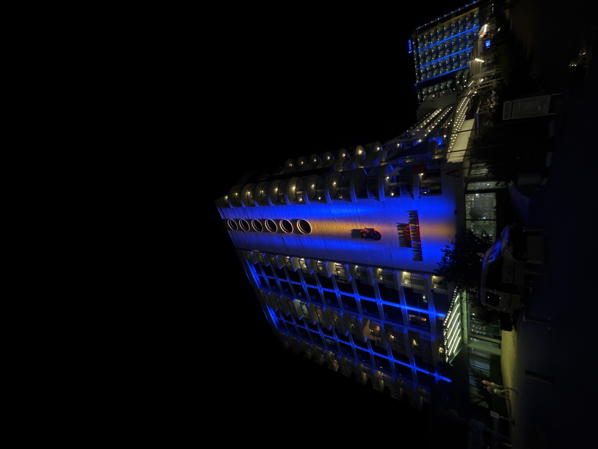 The image shows a night view of the hotel "Berlin Golden Beach" located in Varna, Bulgaria. The hotel is illuminated with blue and yellow lights. The facade of the main building is a tall, curved structure with multiple balconies, lit by blue vertical light strips. A prominent vertical column of lights, transitioning from blue to yellow, runs up the side of the building, highlighting circular decorative elements at the top. A smaller, adjacent building also displays blue lights on its facade in a grid pattern. In the foreground, there is a building entrance with an illuminated canopy. A white van with its side door open is parked in front of the entrance, and a person is partially visible near the van. Other street furniture, including posts and a sign, are present. The scene is dark, with the exception of the artificial lighting from the hotel and surrounding structures.
