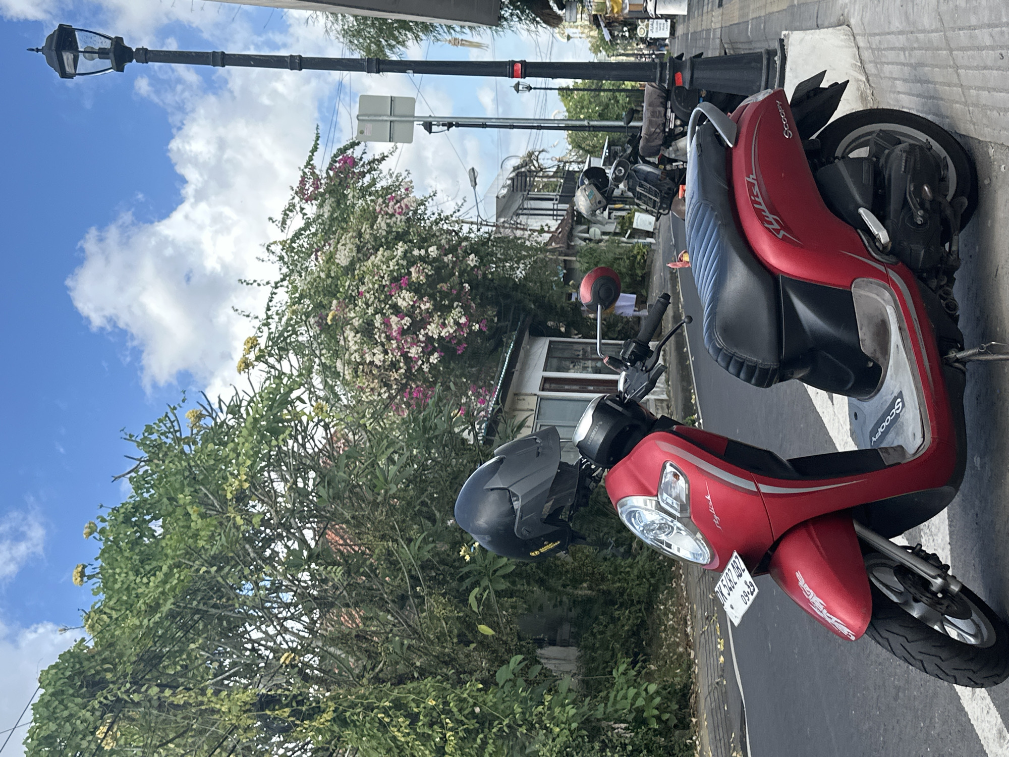 A bright red scooter is parked on the side of a street in Ubud, Indonesia, with its front wheel pointed towards the viewer. A black helmet is resting on the handlebars. In the background, lush green trees and flowering bushes with pink and white blossoms create a vibrant natural scene. A traditional street lamp with a dark metal finish stands tall to the left of the scooter, against a backdrop of a blue sky with fluffy white clouds. The scene suggests a warm, sunny day, typical of a tropical location. The scooter has a license plate with the text "DK 5482 ABZ" and "09-29". The word "Scoopy" is visible on the side of the scooter.