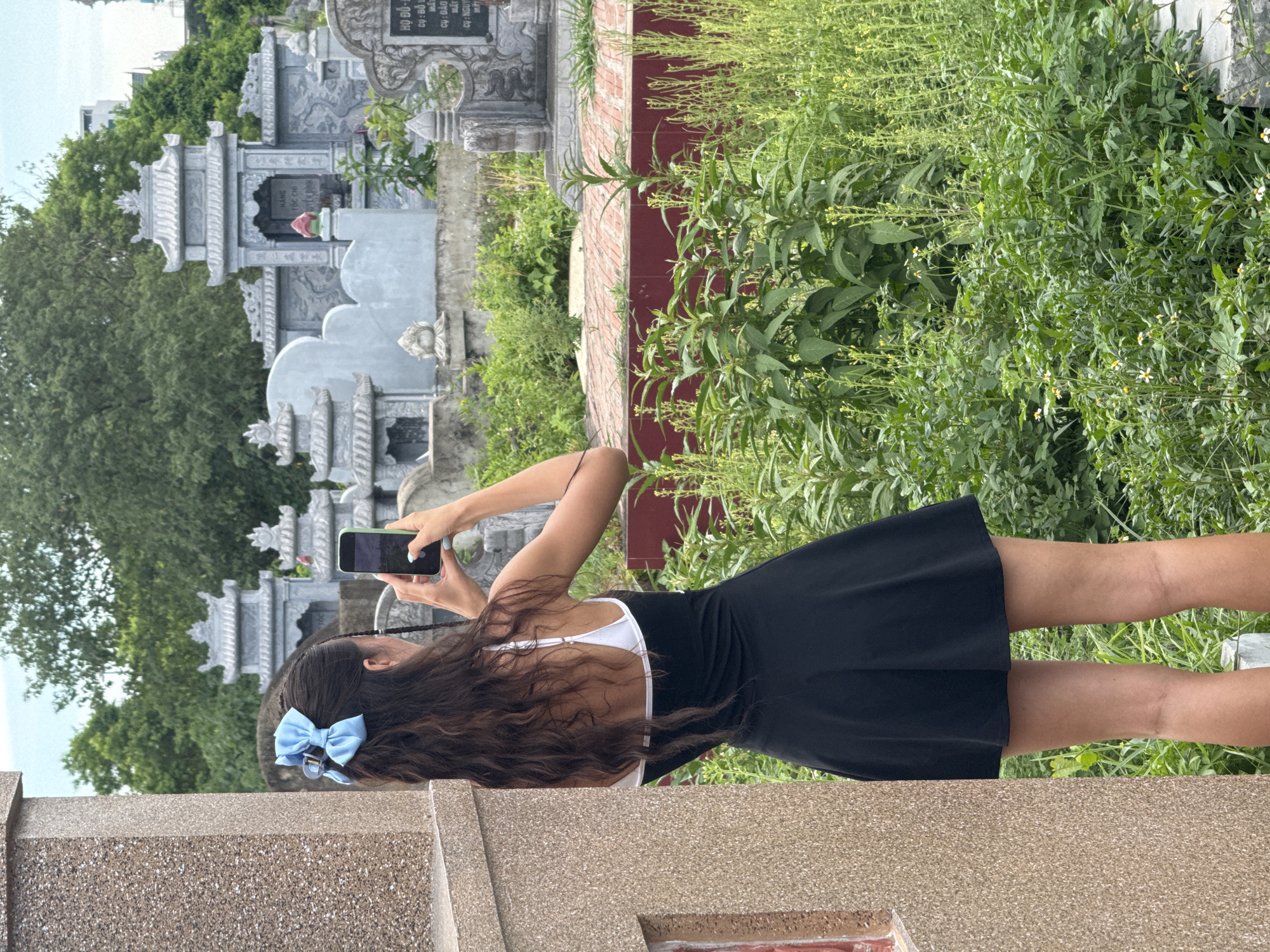 A young woman with long, dark, wavy hair, adorned with a light blue bow, stands in a graveyard in Hoa Lư Ward, Vietnam. She is viewed from behind, wearing a black sleeveless dress with a white trim and a white bralette. The woman is holding a smartphone in her right hand, angled to take a photograph. She is positioned next to a tan, rough-textured wall on the left side of the frame. In the background, numerous grey, ornate gravestones and structures, characteristic of Vietnamese cemeteries, are visible amidst lush green vegetation and trees under an overcast sky. The ground in the immediate vicinity of the gravestones appears to be paved with reddish-brown bricks.