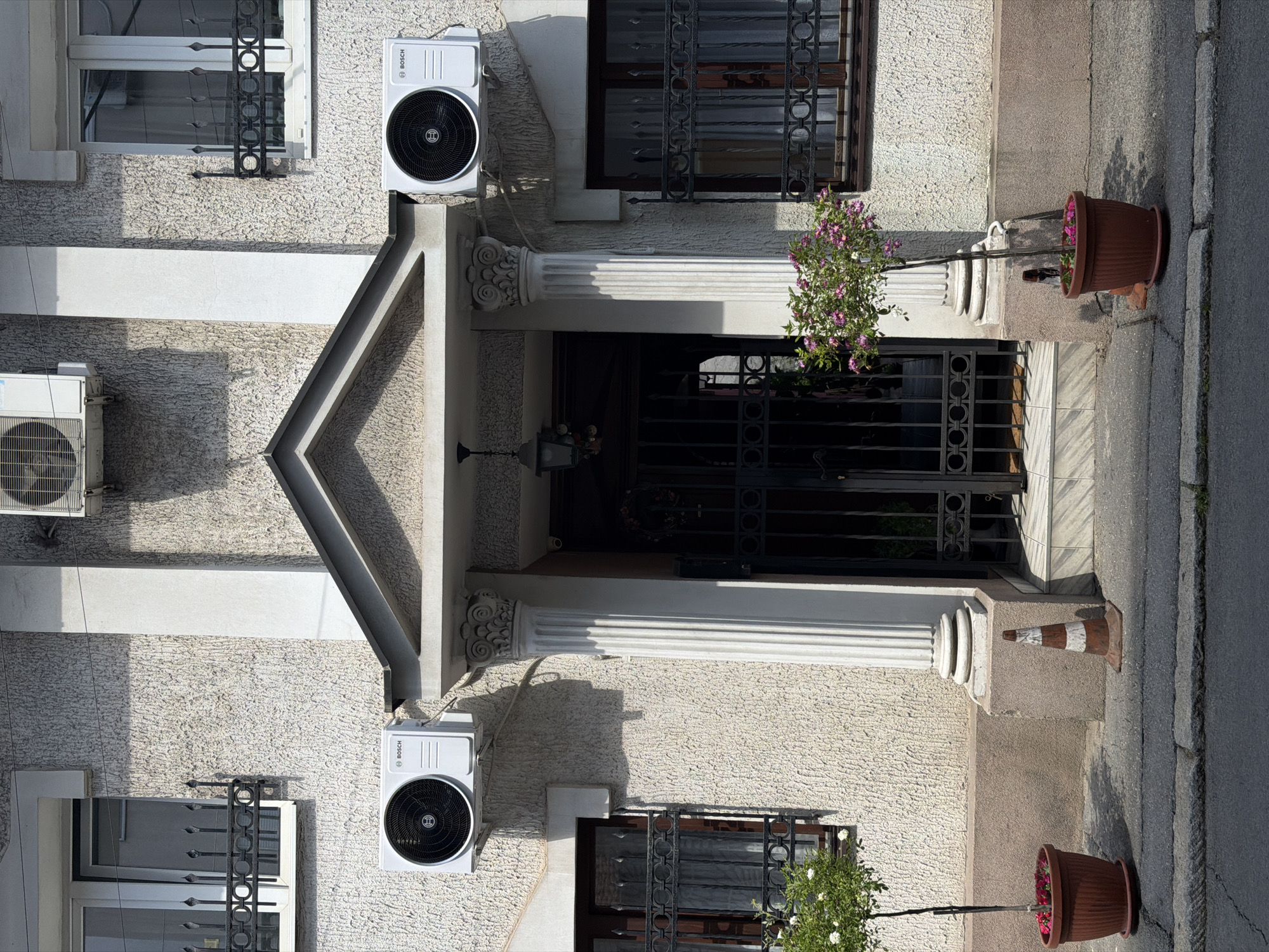 The image shows a building facade in Belgrade, Serbia, with a main entrance framed by ornate columns. The building is light-colored with a rough textured surface. There are windows on either side of the entrance with decorative metal bars. Air conditioning units are mounted on the wall near the windows. Flower pots sit on the ground near the entrance, and one side has a striped traffic cone. The image is taken at an angle, possibly from across the street. The sidewalk is visible on the right side of the frame. The main entrance has black gates.