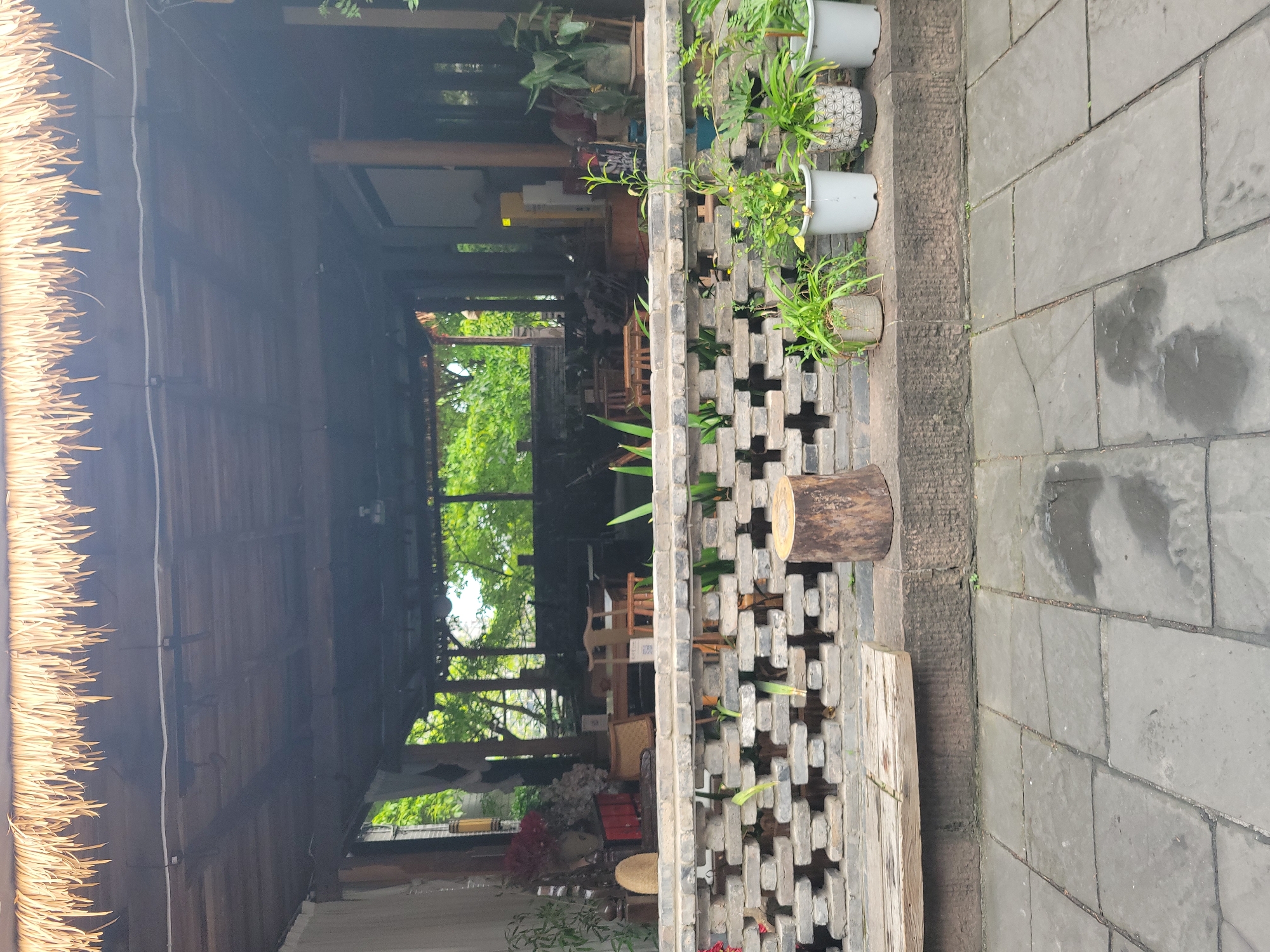 The image depicts a view through a building towards a green area, likely a courtyard or garden. The building has a thatched roof on the left side and is open, with a wooden structure. Inside, there are tables and chairs, suggesting it may be a cafe or restaurant. The view outside shows trees and foliage, indicating a natural setting.
To the right, a gray brick wall with a cross-shaped pattern serves as a barrier. Plants are potted on top of the wall, adding greenery to the scene. A segment of a tree trunk rests on the wall, possibly serving as a decorative element or a seat. The ground is paved with large, gray rectangular stones.
While the image is generally well-lit, the interior of the building appears somewhat darker than the exterior. Based on the prompt, the location is in Hechuan, China, though the specific type of buildings and style don't support or contradict this. No people are visible in the frame.