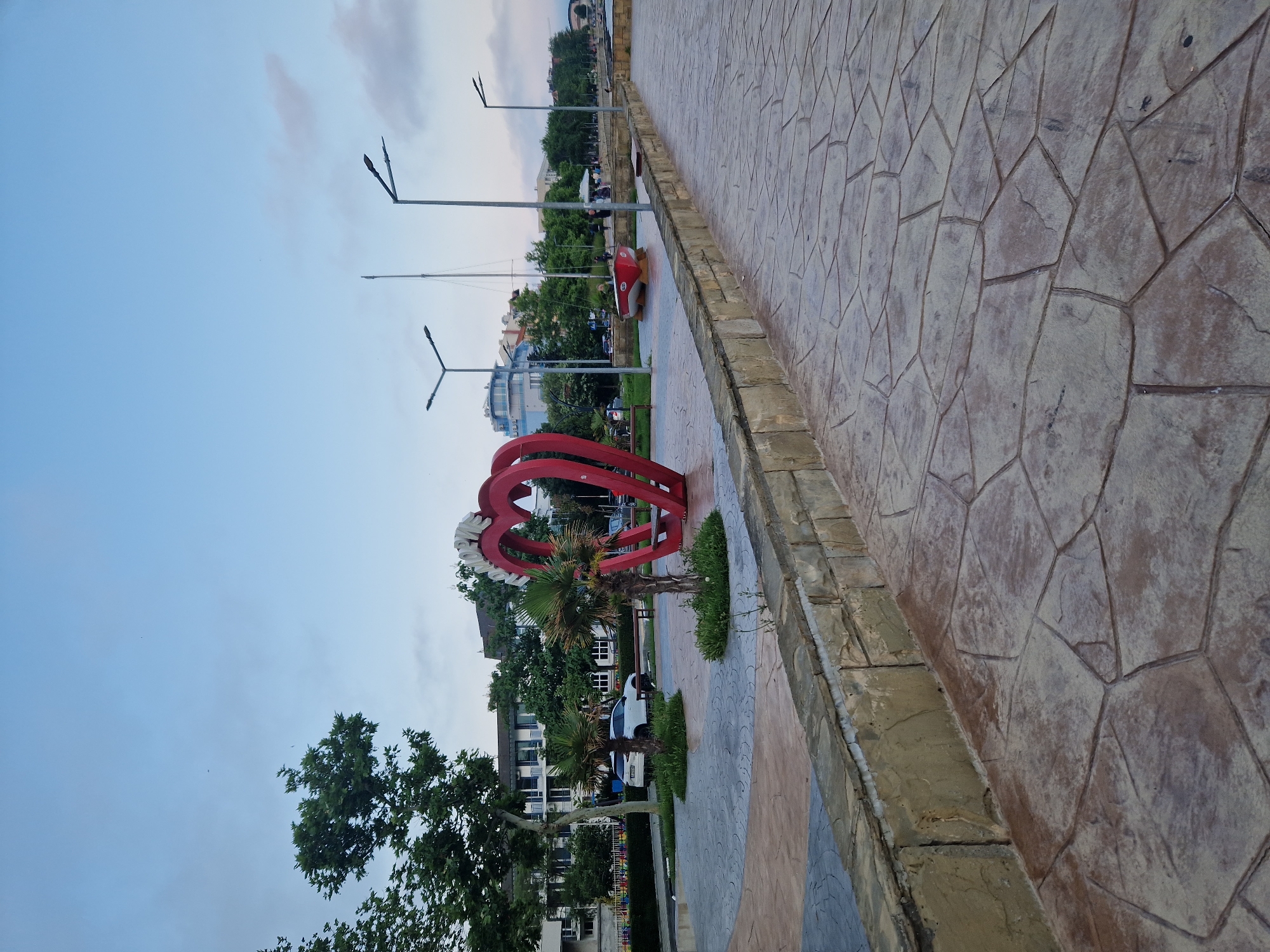 This image depicts a waterfront promenade in Pomorie, Bulgaria, likely during overcast daytime or twilight.
The foreground features a wide pedestrian path paved with irregular, dark brown and grey flagstone-patterned concrete. A low, rough-cut stone wall, varying in color from tan to light brown, separates the path from a landscaped park area.
In the midground, a prominent large red heart-shaped sculpture stands. Cyrillic lettering, 'ПОМОРИЕ,' is visible on its upper right curve. Two small, bushy palm trees are planted in circular beds near the heart. Further left, a large deciduous tree with dense green foliage provides shade. A white SUV is parked behind the heart sculpture on an adjacent street.
To the right of the heart, a small, red-hulled boat with a white bottom is dry-docked on a grassy patch. Multiple modern streetlights with twin angled luminaires are positioned along the promenade and the street. In the background, several multi-story buildings are visible; one on the left is white with balconies, and another to the right is light blue with a curved facade. A few indistinct figures of people are present in the distant midground near the boat and further down the promenade.
The sky is a pale blue with scattered, thin, light grey clouds, and a few small, dark specks, possibly birds, are discernible in the upper left quadrant.