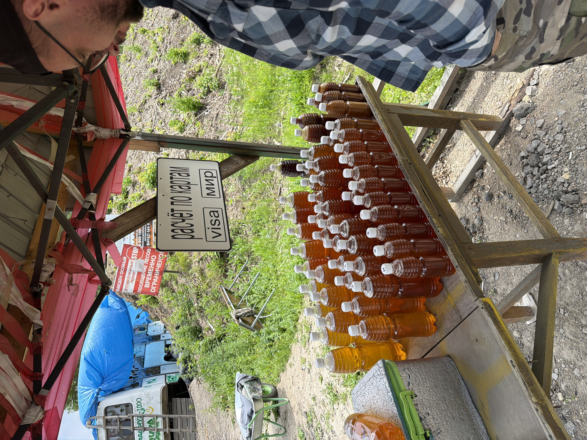 In Tarmanchukan, Russia, a roadside stall displays rows of plastic bottles filled with amber-colored liquid, presumably honey. The stall is covered by a red and white striped canopy, partially collapsed. A sign above the bottles reads "расчёт по картам" (payment by card), with "visa" and "мир" (a Russian payment system) indicated below. A person wearing glasses and a plaid shirt stands to the right, looking down at the stall. In the background, a white bus covered in a blue tarp is parked, and a wheelbarrow rests on the gravelly ground. Green vegetation grows around the stall and the bus.