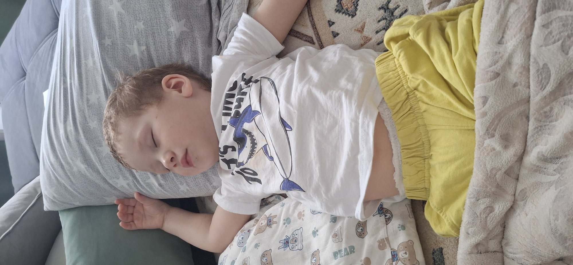 A young child, likely a boy, is seen sleeping peacefully in an indoor setting. The child is lying on their left side, with eyes closed and a calm expression, resting their head on a light gray pillow patterned with subtle white stars. Their hair is light brown and appears slightly tousled.
The child is wearing a white short-sleeved t-shirt featuring a blue and white shark graphic on the front, along with some partially visible text, possibly "SHARK" or related. Below the t-shirt, light-colored shorts or pajama bottoms are visible, adorned with various cartoon bear faces and paw prints, including the word "BEAR" printed on them.
The immediate surroundings suggest a comfortable sleeping area. To the child's left, a dark gray sofa or bed is partially visible, along with a sage green cushion. To the child's right, a bright yellow garment or blanket is crumpled, and further right, a patterned beige or light brown rug or blanket with an intricate design is seen, overlaid with a white textured blanket. The overall lighting is soft and natural, suggesting it is daytime. The scene evokes a sense of tranquility and rest.