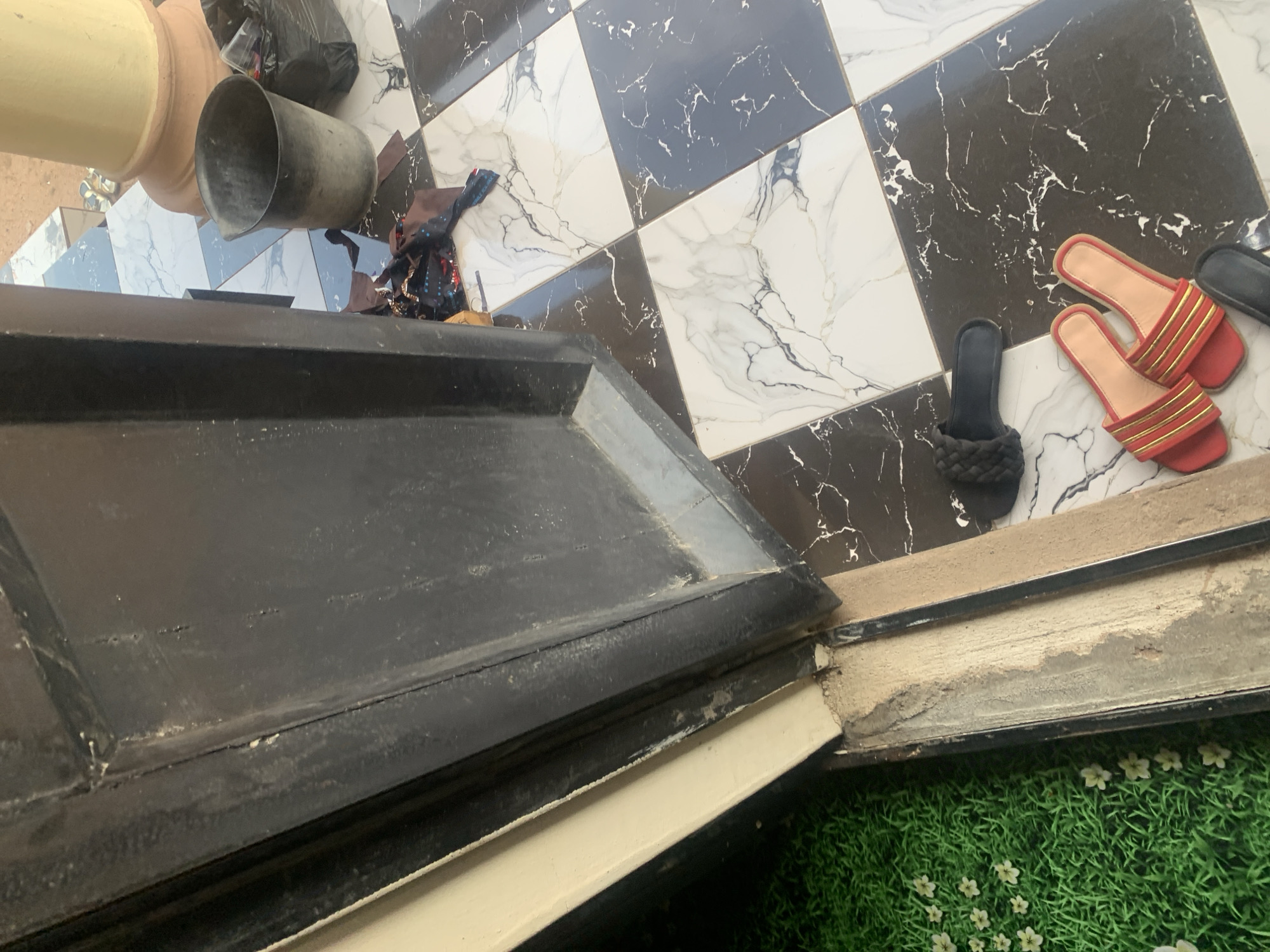 The image depicts an outdoor scene in Jauro Yinu, Nigeria, featuring a doorway with black-painted wood. The ground is tiled with a pattern of black and white marble squares. Several objects are scattered on the tiles. In the foreground, a pair of red and gold slides is visible. To the left of the slides, a black slipper with a woven strap is present. Near the doorway, a black plastic bucket and a black garbage bag filled with items are situated. A piece of patterned fabric lies on the floor between the bucket and the door. To the far left, a green mat with a floral pattern of white flowers can be seen. A cylindrical, cream-colored pillar is partially visible in the background on the right side of the frame.