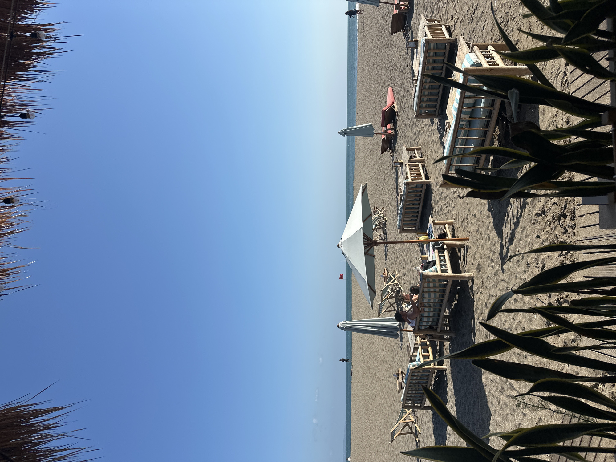 The image depicts a serene beach scene in North Kuta, Indonesia, under a clear blue sky. In the foreground, a thatched roof provides shade, with glimpses of hanging lightbulbs. The sandy beach is dotted with various seating arrangements, including wooden benches with striped cushions and lounge chairs. A couple is seated on one of the benches, engaged in conversation. Further back, several umbrellas offer shade on the beach, with two red lounge chairs set up near one. In the distance, the calm ocean stretches towards the horizon, where a lone figure stands near the water. A small red flag is visible on a pole in the mid-ground. The overall impression is one of tranquility and relaxation, characteristic of a beach resort.