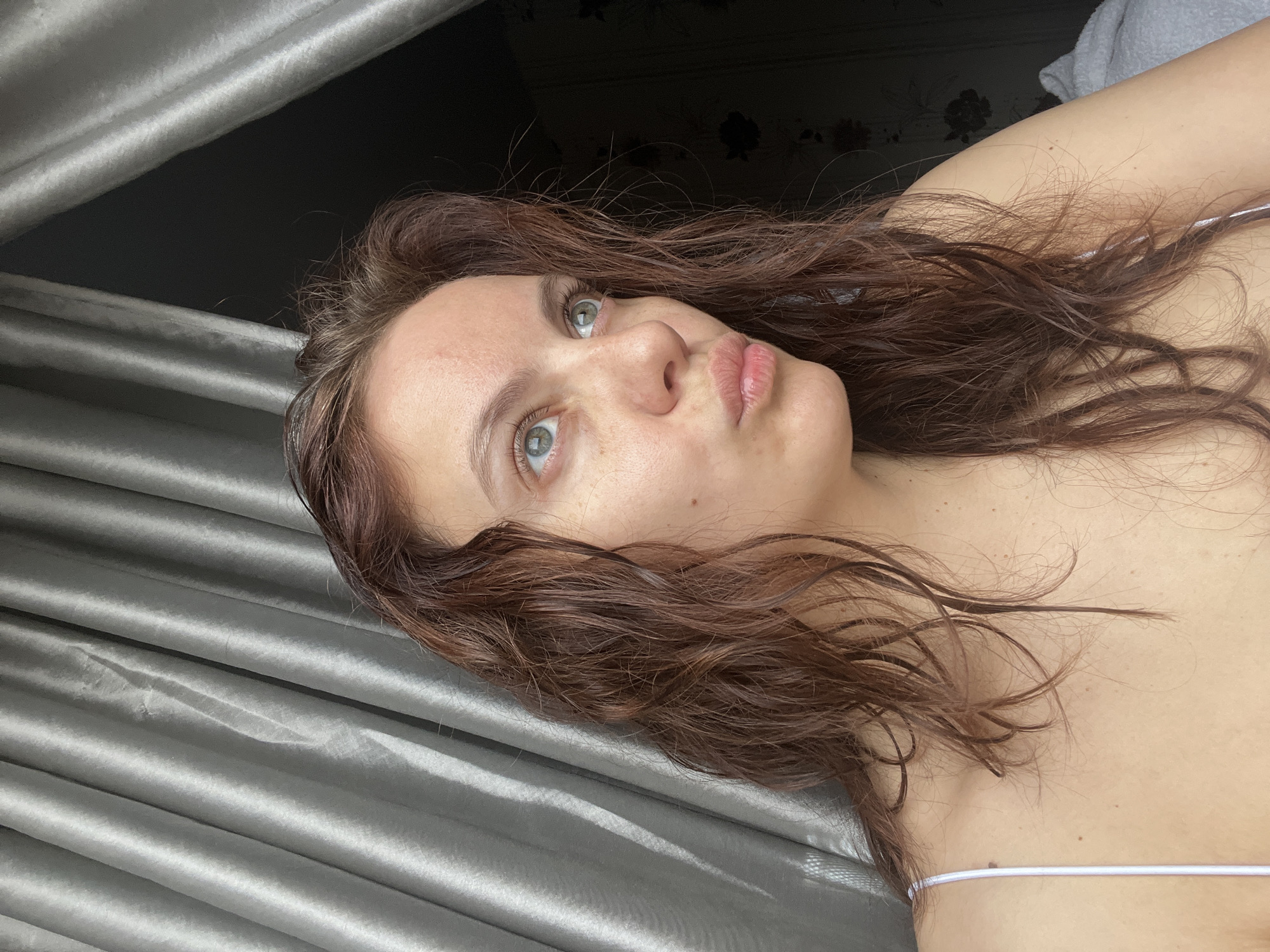 A woman with brown hair and green eyes is looking up and to the left with a slightly pursed expression. Her hair is wet and appears to be wavy. She is wearing a white strap. The background consists of light gray, vertically draped fabric to the left and a dark surface to the right. The city and country are not discernible from this image.