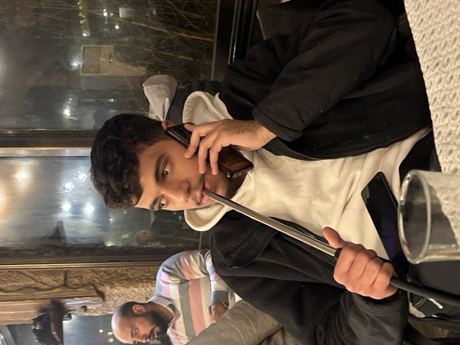 A young man in a black jacket over a white hoodie smokes a hookah while talking on a cell phone. He looks directly at the camera. His other hand holds the hookah hose. To his left, an older man sits with a striped shirt. A second phone and glass are on the table. A window with blurred city lights is visible in the background.