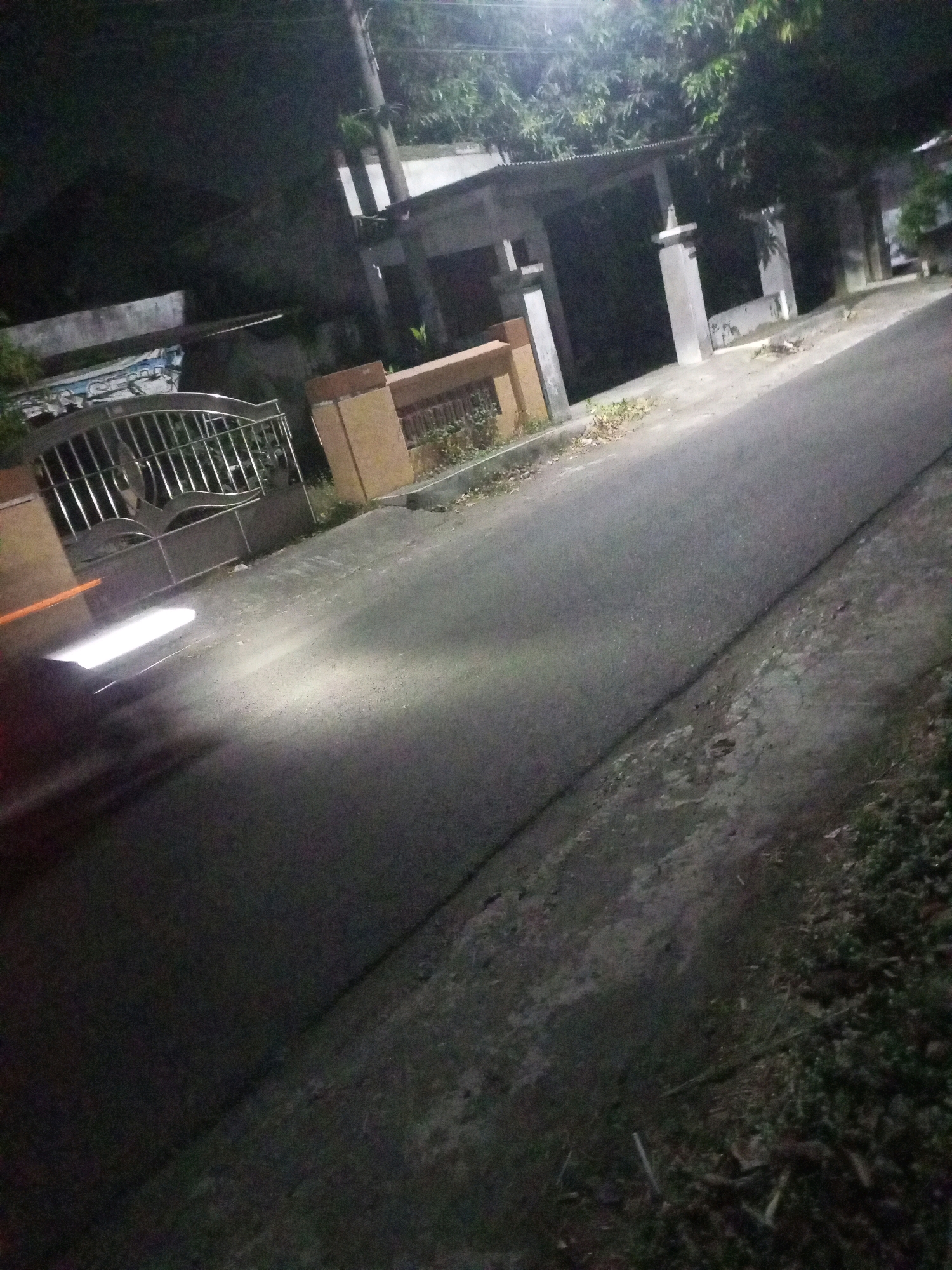 In Ngasem, Indonesia, at night, a dimly lit asphalt road runs diagonally from the bottom right to the top left of the frame. The road surface shows signs of wear, with patches of concrete and dirt. To the left, a metal gate with intricate designs separates the road from a property. A bright white light, likely from a vehicle, illuminates a section of the road in the lower left corner, creating a blurred streak of light and motion. Beyond the gate, a building with a corrugated roof and concrete pillars is visible, partially obscured by darkness and foliage. The overall scene suggests a residential or semi-urban area at rest under the cloak of night.