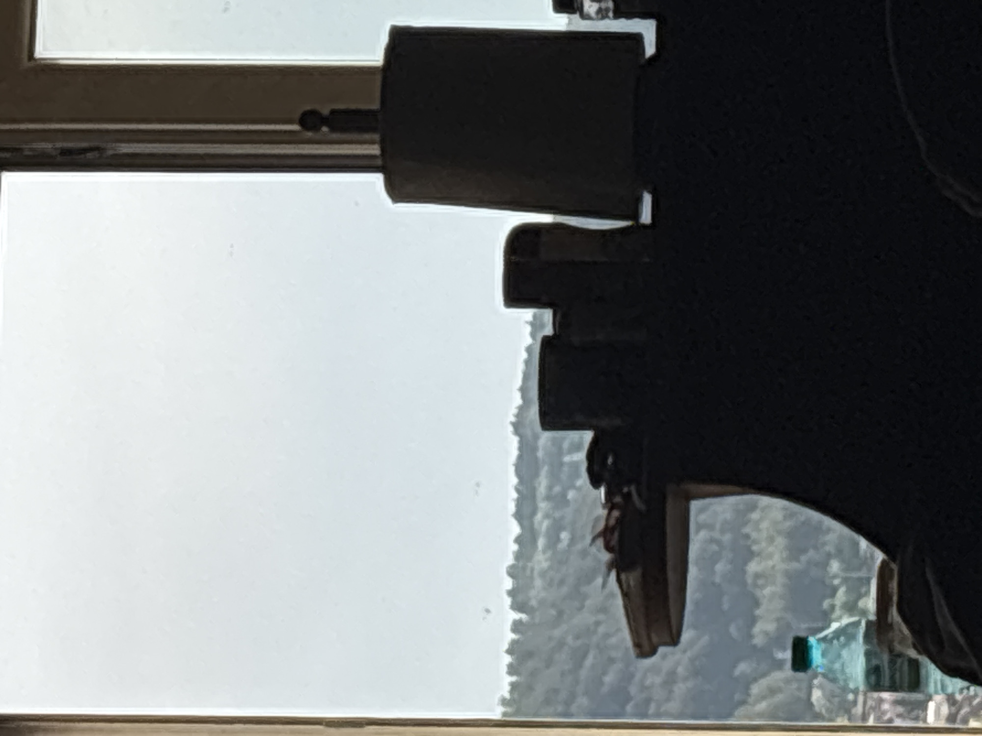 The image shows a view from inside a building, possibly a home, looking out through a window. The window has a light-colored frame. Outside, there is a hazy view of a landscape, likely a forest or hills covered in trees. In the foreground, partially obscuring the view, is a black object, possibly part of a camera or other equipment. A water bottle is visible in the lower right corner. The location is unknown.