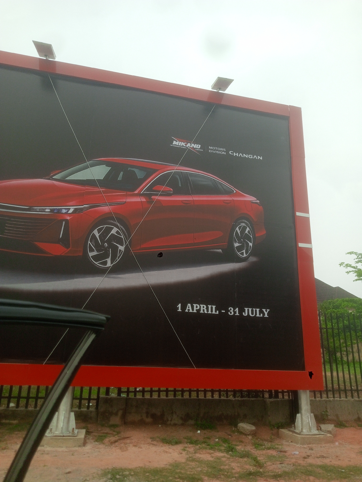The image shows a large outdoor advertisement in Kubwa, Nigeria. The advertisement is for Changan Motors, specifically featuring a red car. The advertisement includes the text "1 APRIL - 31 JULY". The billboard has a red frame. There are two lights on the top of the billboard. The billboard is situated next to a fence and vegetation. The sky is overcast. A car doorframe is visible on the left side of the image, partially obscuring the view.