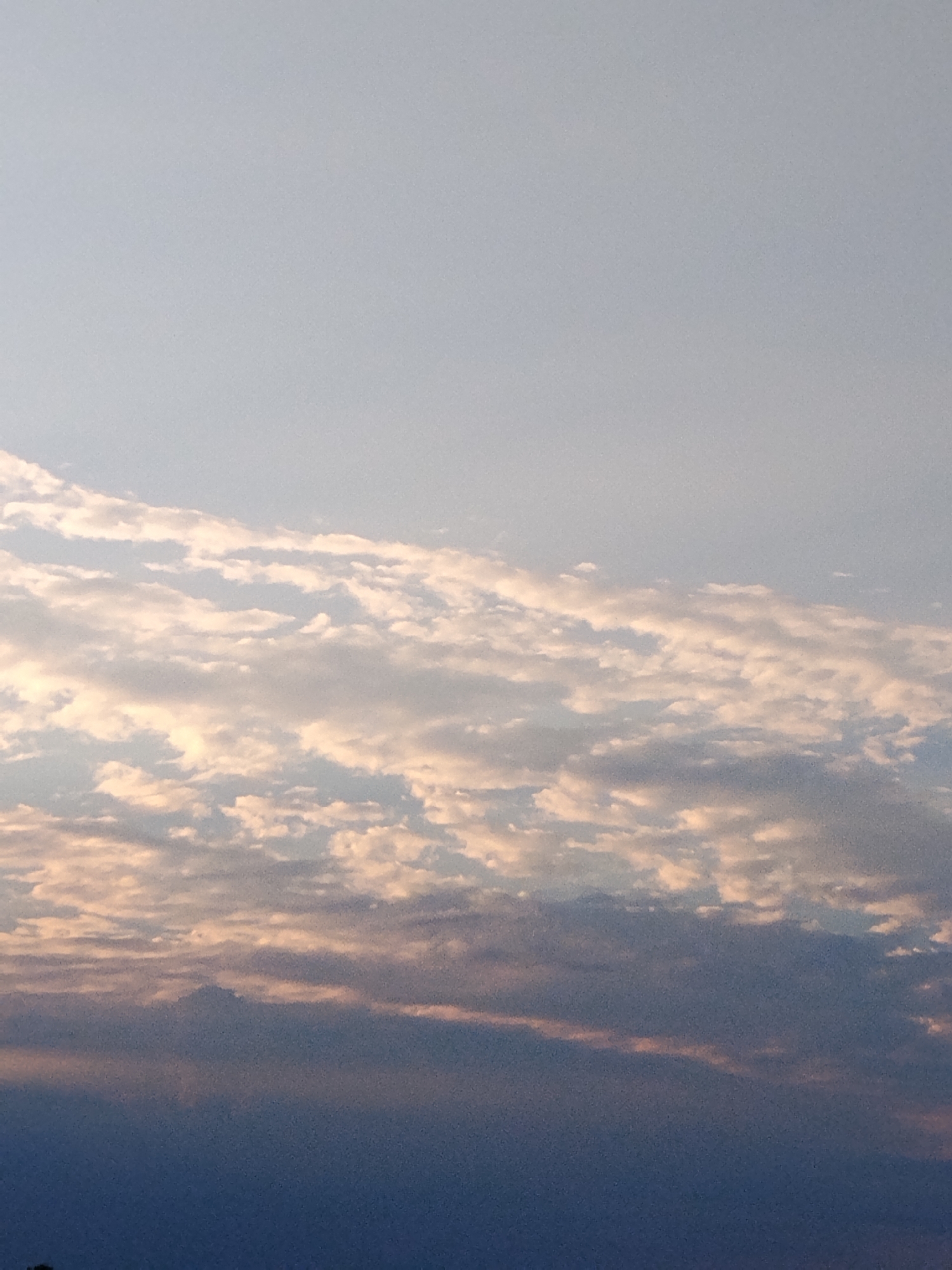 The image shows a wide shot of a sky filled with clouds. The sky is a light blue, and the clouds are a mix of white and gray, with some hints of orange and pink, suggesting it is either sunrise or sunset. The clouds are scattered across the sky in various sizes and formations, with some appearing wispy and others more dense. The bottom portion of the image is darker, possibly indicating the silhouette of the ground or distant objects against the bright sky. There is no visible text in the image. The overall mood of the image is serene and peaceful, characteristic of a natural landscape at twilight.