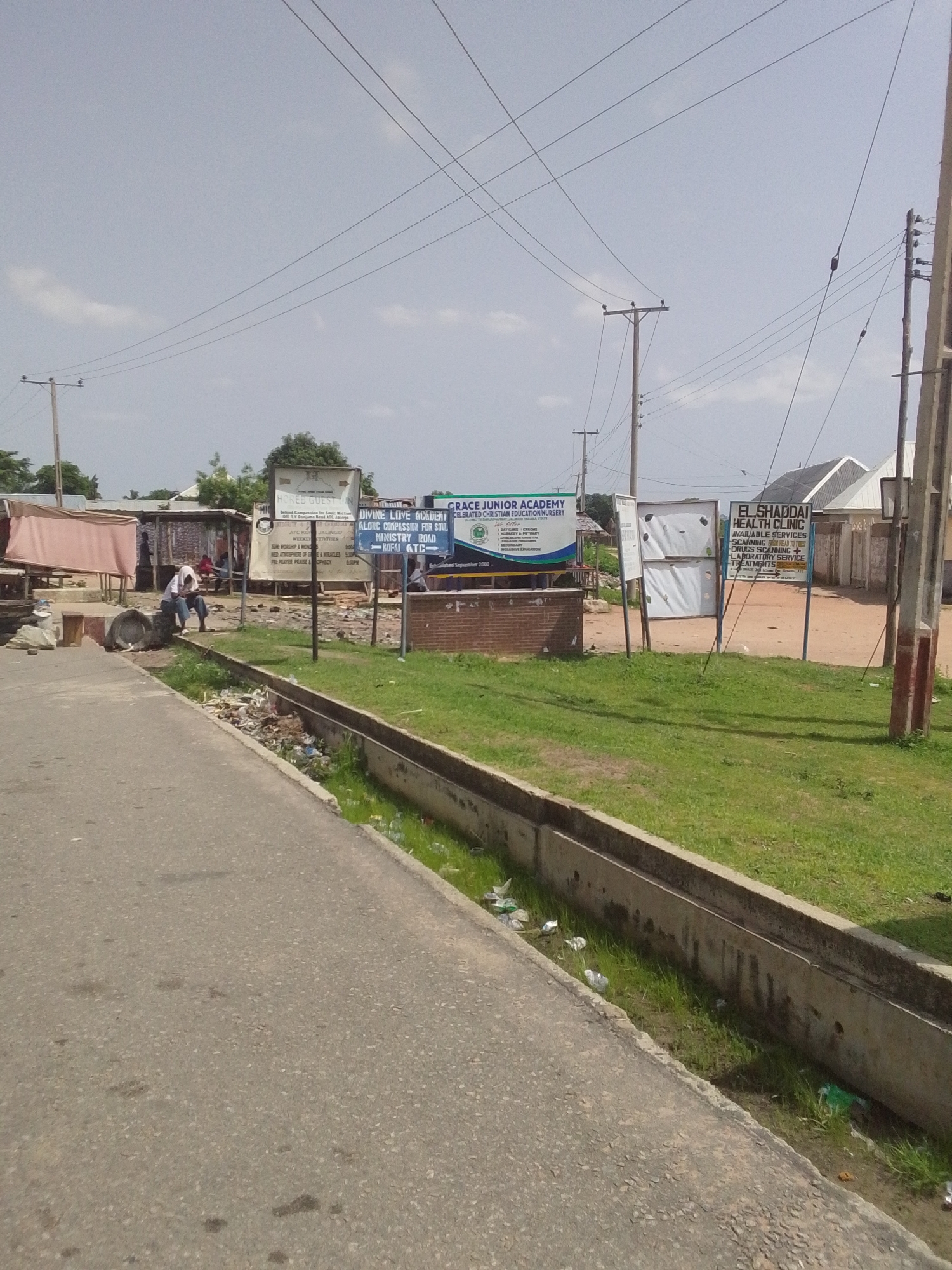 This media file captures an outdoor scene likely in Jalingo, Nigeria. The foreground shows a paved road with a concrete drainage ditch running alongside it. The ditch contains some grass and scattered litter, suggesting it may not be well-maintained.
The midground reveals a street with various businesses and signs. To the left, there are some open-air stalls or shops with covered awnings, where a few people are visible, one sitting on a concrete structure. Further to the right, there are multiple signs for educational and health institutions. One sign reads "GRACE JUNIOR ACADEMY" and "DIVINE LOVE ANTLERY CELERATED CHRISTIAN EDUCATION NURSERY," and another indicates "EL SHADDAI HEALTH CLINIC" with "AVAILABLE SERVICES" like scanning, drugs, and laboratory services. There are also two large, blank white boards on poles.
The background shows more buildings, some with pitched roofs, and a cluster of trees. Utility poles with overhead wires crisscross the sky, indicating the presence of electricity infrastructure. The sky is mostly clear with a few faint clouds, suggesting it is daytime. The overall atmosphere appears somewhat dusty and perhaps not a highly developed urban area, given the visible litter and basic structures. The presence of the signs suggests that these are functioning establishments, implying a busy community.