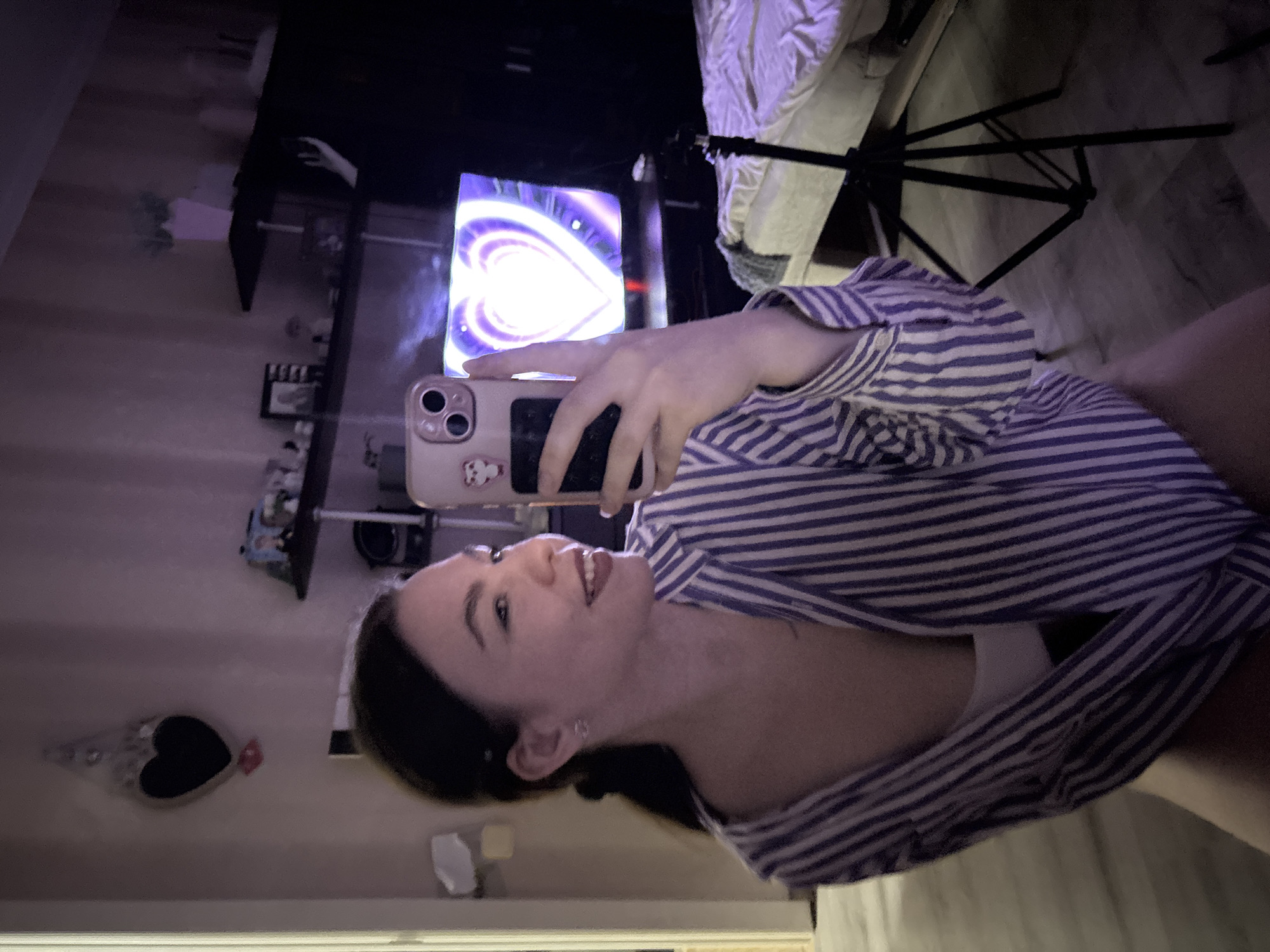 This media file features a young woman taking a mirror selfie indoors, likely in Perm, Russia. She is positioned in the foreground, facing towards the left of the frame, with a pleasant, relaxed smile on her face and looking slightly upwards. Her dark hair is visible, and she is wearing a blue and white striped button-down shirt over a white undershirt. She holds a light-colored smartphone with a distinctive camera module and a small, light-colored cartoon sticker (resembling a bear or dog) on its back.
The setting appears to be a room, possibly a bedroom, captured during the evening or night due to the prominent artificial lighting. In the background, a large screen (possibly a television or monitor) displays a vibrant, glowing purple and white heart graphic with circular patterns emanating from its center. Below the screen, dark shelves or furniture are visible, holding several small, indistinct objects. To the right of the screen, a black tripod stand, commonly used for lighting equipment like a ring light or softbox, stands next to what appears to be a white piece of furniture or bedding. On the left wall, a dark, heart-shaped decorative item is hanging. The floor is light-colored. The overall lighting scheme gives the image a cool, slightly purple hue, contributing to a cozy and personal atmosphere. There is no visible text in the image.