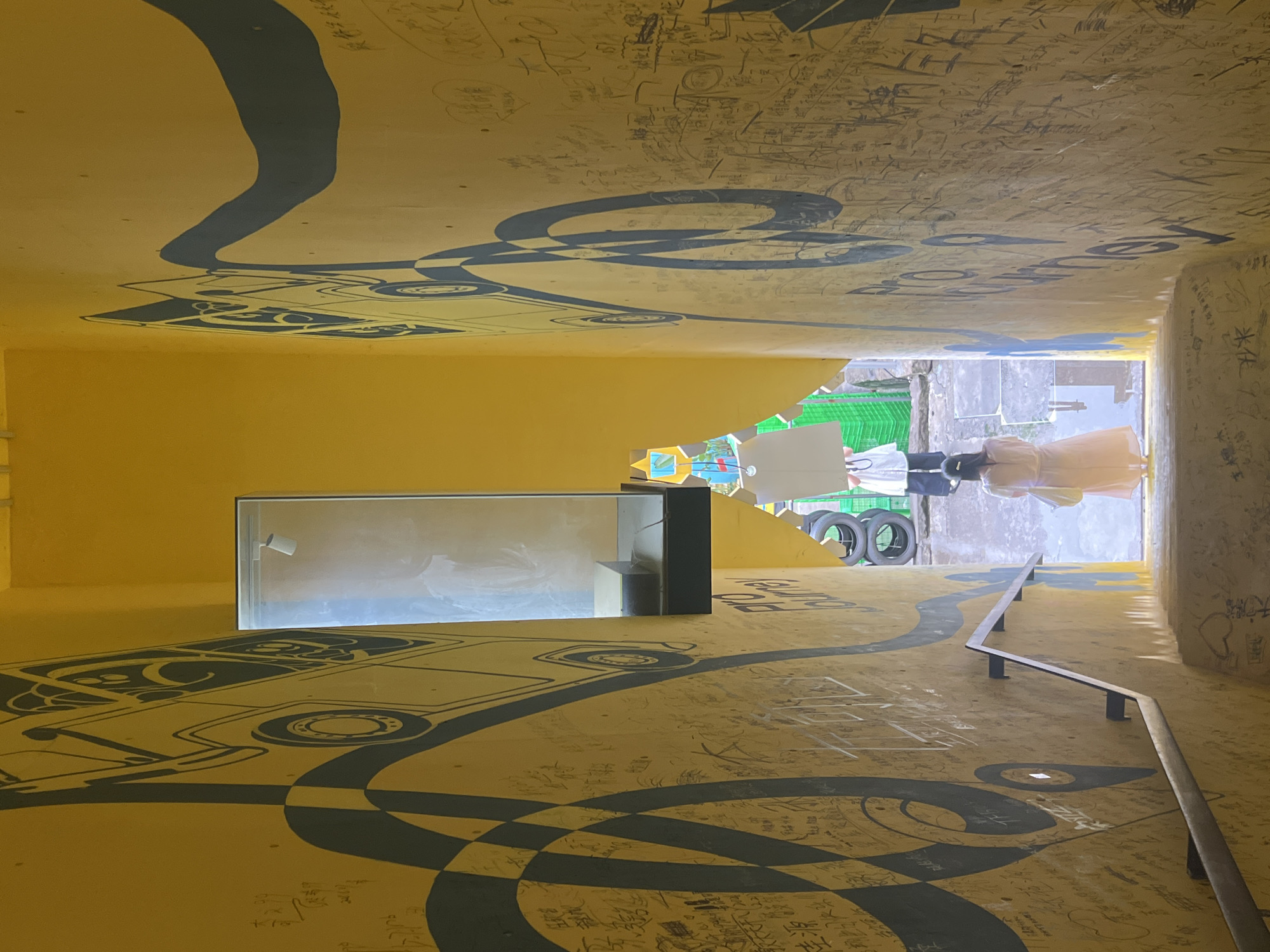 This image shows a brightly lit yellow staircase with abstract black murals on the walls, depicting stylized vehicles and curving lines. The walls are covered in numerous handwritten inscriptions, suggesting a public space where people have left their mark.
A metal handrail runs along the left side of the staircase, providing support for descending. At the top of the staircase, the path opens to an outdoor area. Two individuals are visible at the top of the stairs, facing away from the camera. One person, wearing a white top and dark pants, appears to be holding a white object. Another person, in a light-colored dress, is positioned further down the stairs, also facing away and carrying a light-colored bag.
In the background of the outdoor area, there are stacks of black tires and green crates, hinting at a utilitarian or possibly an industrial setting. A glass-encased display or niche is embedded in the wall on the right side of the staircase, further up.
The overall impression is of an artistic and interactive urban environment, likely a public walkway or passageway in Yuzhong District, China, where graffiti and artistic elements are integrated with the architecture. The sunlight filtering in creates a strong contrast between the illuminated areas and the shadows cast by the staircase.