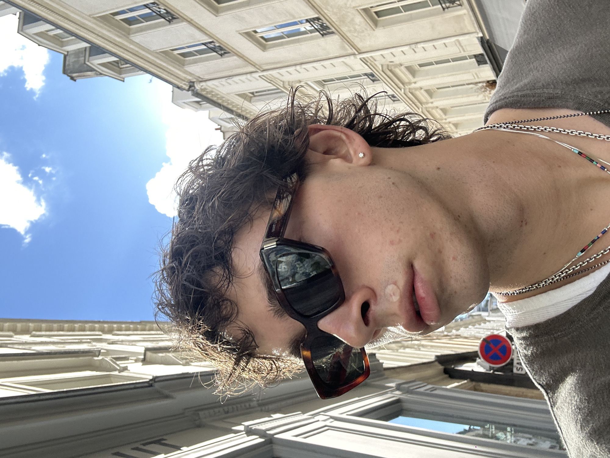 A low-angle shot of a person wearing sunglasses and necklaces standing in Paris, France. The person has short, curly, brown hair and is wearing a gray shirt. They have a clear pimple patch on their face. The buildings of Paris are visible in the background, along with a blue sky and some white clouds. A red and blue no parking sign is also visible.