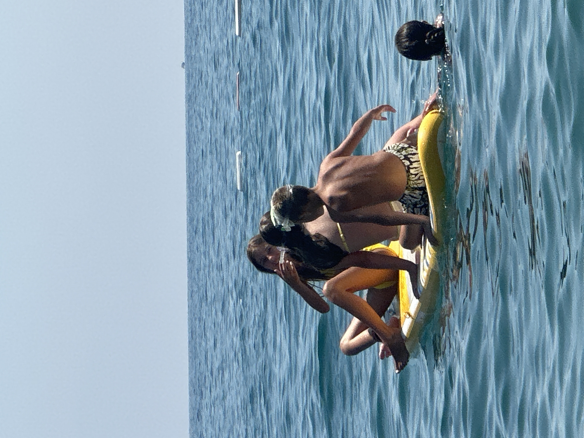 The image depicts a scene at a beach in Boreti, Montenegro. Four children are on a yellow inflatable paddleboard in the calm, blue sea. One girl, wearing a yellow bikini, is seated and appears to be holding a piece of food to her mouth. Another girl, with dark hair and a gold bikini strap visible, is seated behind her. A boy, wearing patterned swim trunks, is sitting on the paddleboard facing away from the camera, and another child's head, with dark hair in a braid, is visible in the water to the right of the paddleboard. The sky is clear and light blue, and a distant boat can be seen on the horizon. There are several white buoys floating in the water.
