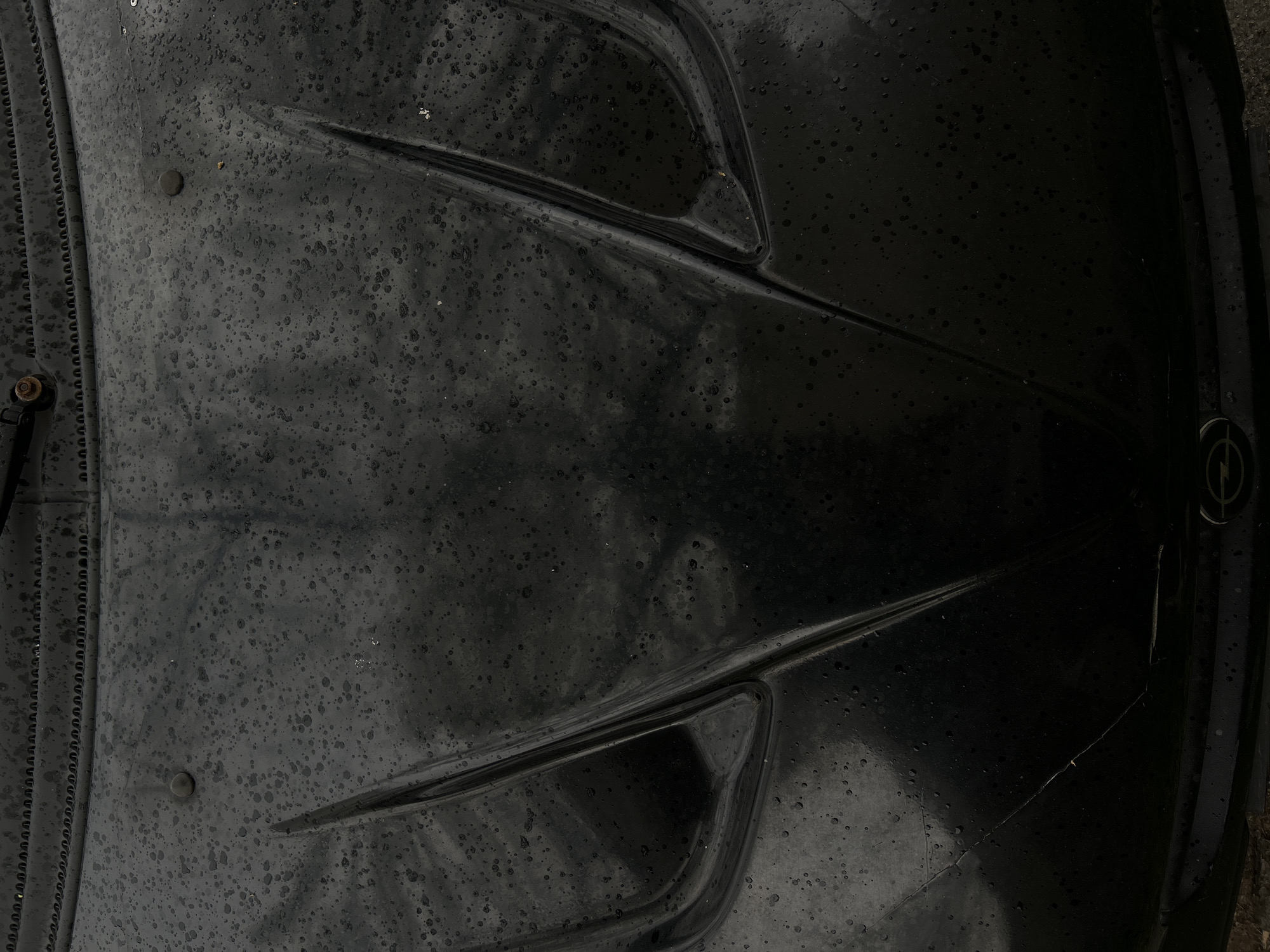 The image shows the front hood of a black Opel car, likely parked outdoors. The hood has two prominent creases running towards the front. The Opel logo is visible on the right side of the hood. The surface is covered in water droplets. Details on the location in Plovdiv, Bulgaria, are not discernable from the image.