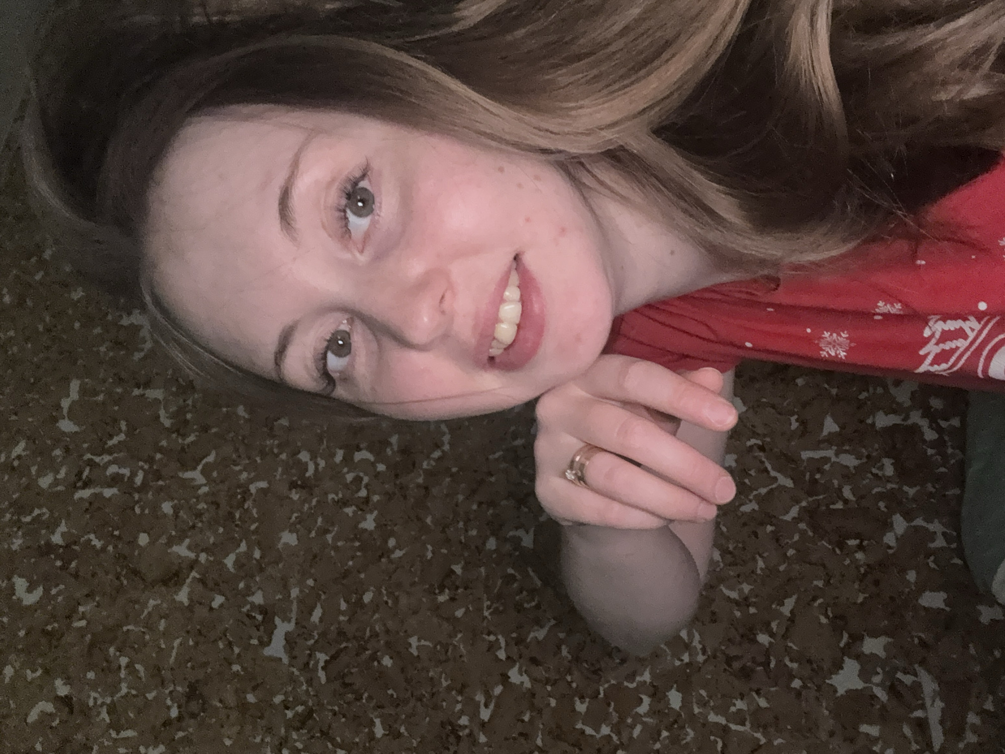 A woman lies on a speckled brown surface in Unknown, Unknown. She is wearing a red shirt with white designs and a gold ring on her finger. Her hair is blonde with brown roots, and she is smiling.