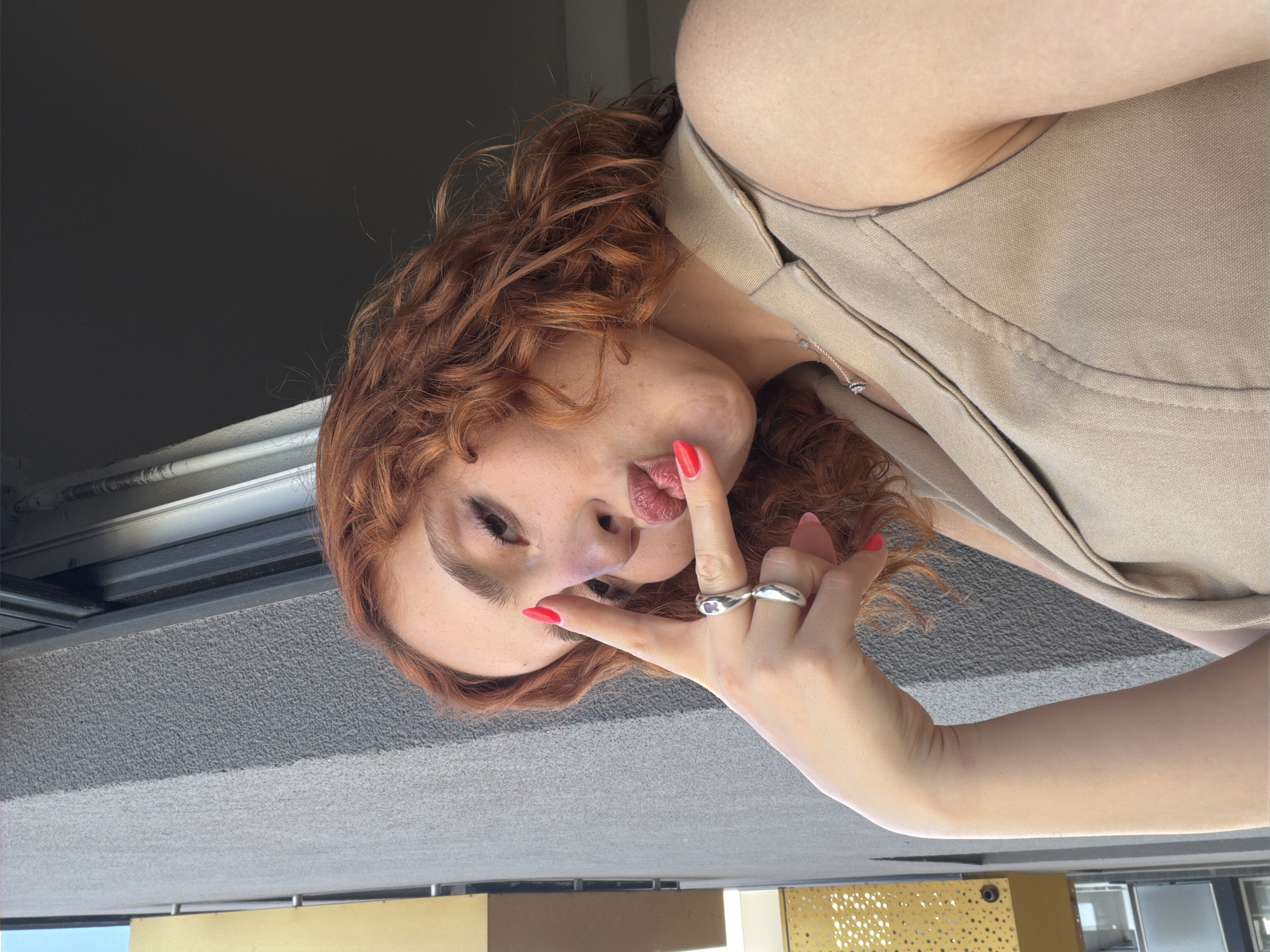A young woman with reddish-brown curly hair and fair skin is taking a selfie outdoors. She is wearing a sleeveless beige vest and a silver necklace with a small diamond pendant. Her left hand is raised to her face, with her index and middle fingers forming a peace sign. She has bright red nail polish on her fingernails and her lips are painted with a similar shade. She is puckering her lips. The background shows a gray, textured wall and a window frame. There is a glimpse of a yellow building on the left side of the frame. The image appears to be taken from a balcony or an elevated position, suggesting an urban environment. The presence of buildings and a balcony indicates a city setting, possibly in Strasbourg, France, given the architectural style.