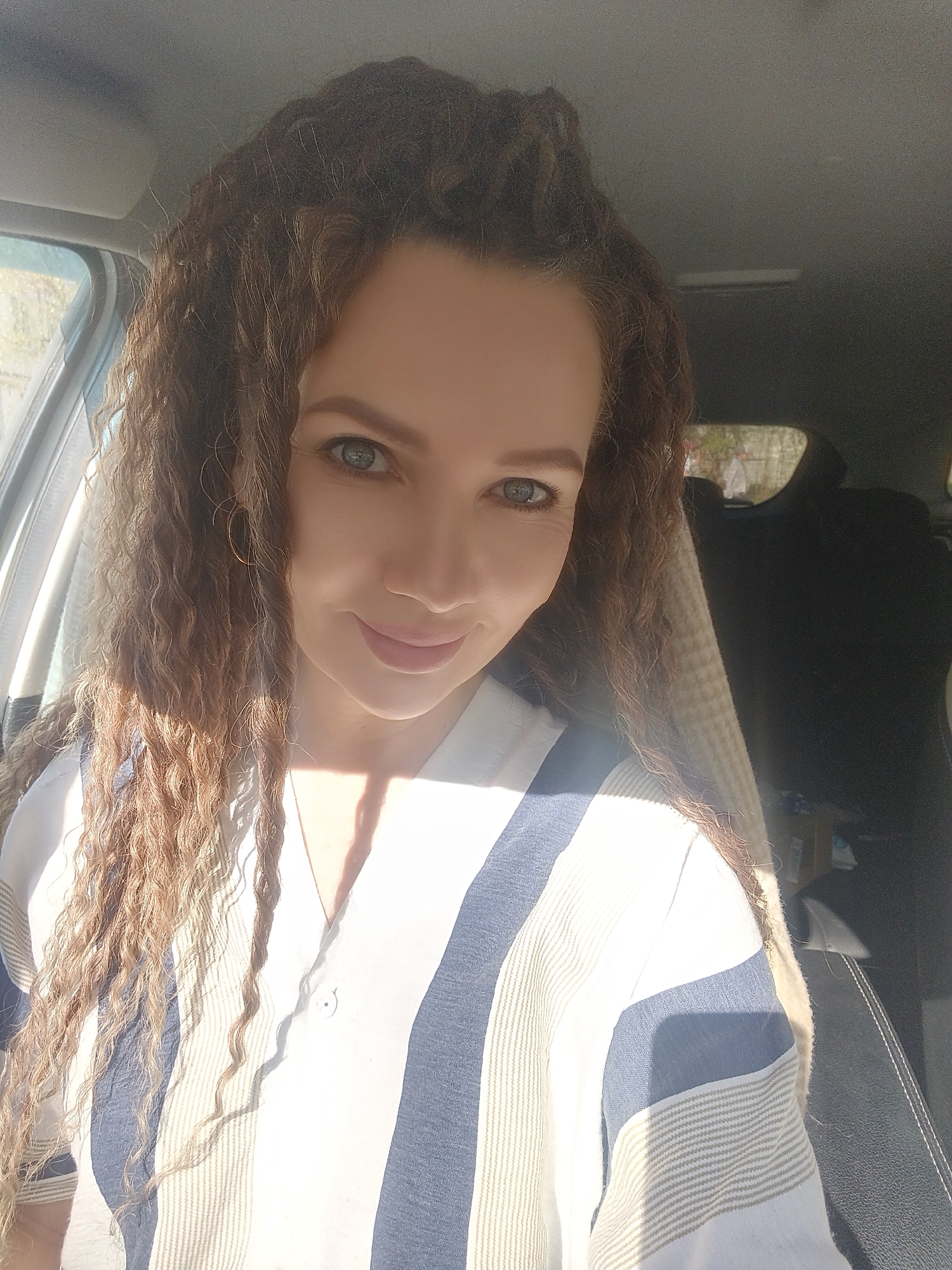 A portrait captures a woman with curly brown hair and light eyes, wearing a striped blue and white shirt. The image is taken inside a car. The location is Unknown, Unknown.