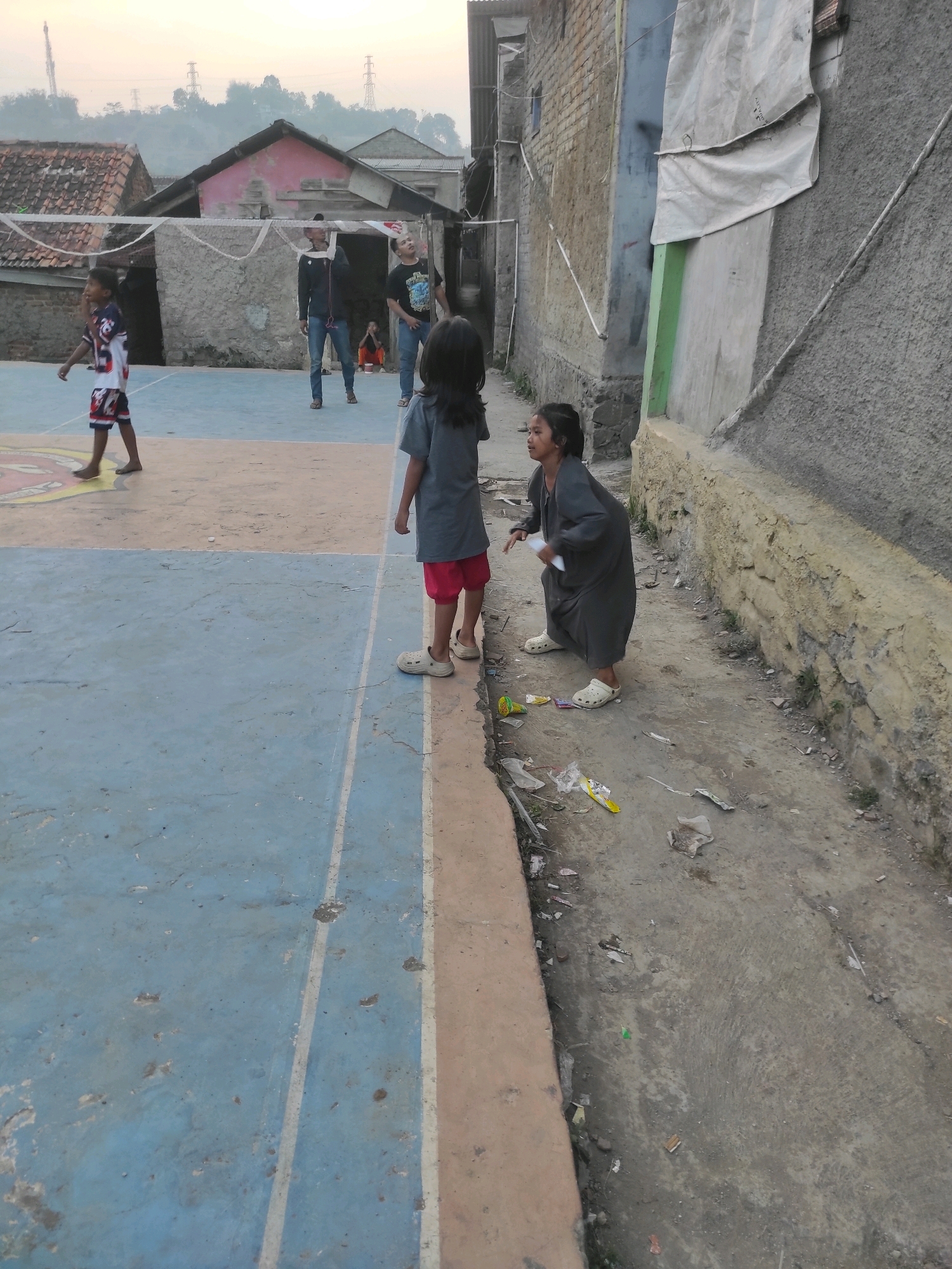 In Padalarang, Indonesia, a basketball court with faded blue and orange markings serves as the backdrop for a casual gathering. A young boy in a patterned jersey and red shorts stands on the court, looking towards the left. Two young girls are positioned near the edge of the court. One, in a gray t-shirt and red shorts, stands facing the other. The second girl, in a dark gray dress and white clogs, is crouched down, looking upwards and to the right, holding a white object. In the background, three men are visible on the court, one in a dark jacket and jeans, another in a black t-shirt and jeans, and a third man partially obscured. A volleyball net stretches across the court. The surroundings show a mix of brick buildings, some with rough plaster, and corrugated metal roofs. The ground near the court is a dirt path littered with various pieces of trash, including plastic wrappers and debris. Hills with vegetation and distant power lines are visible on the horizon under a pale sky.